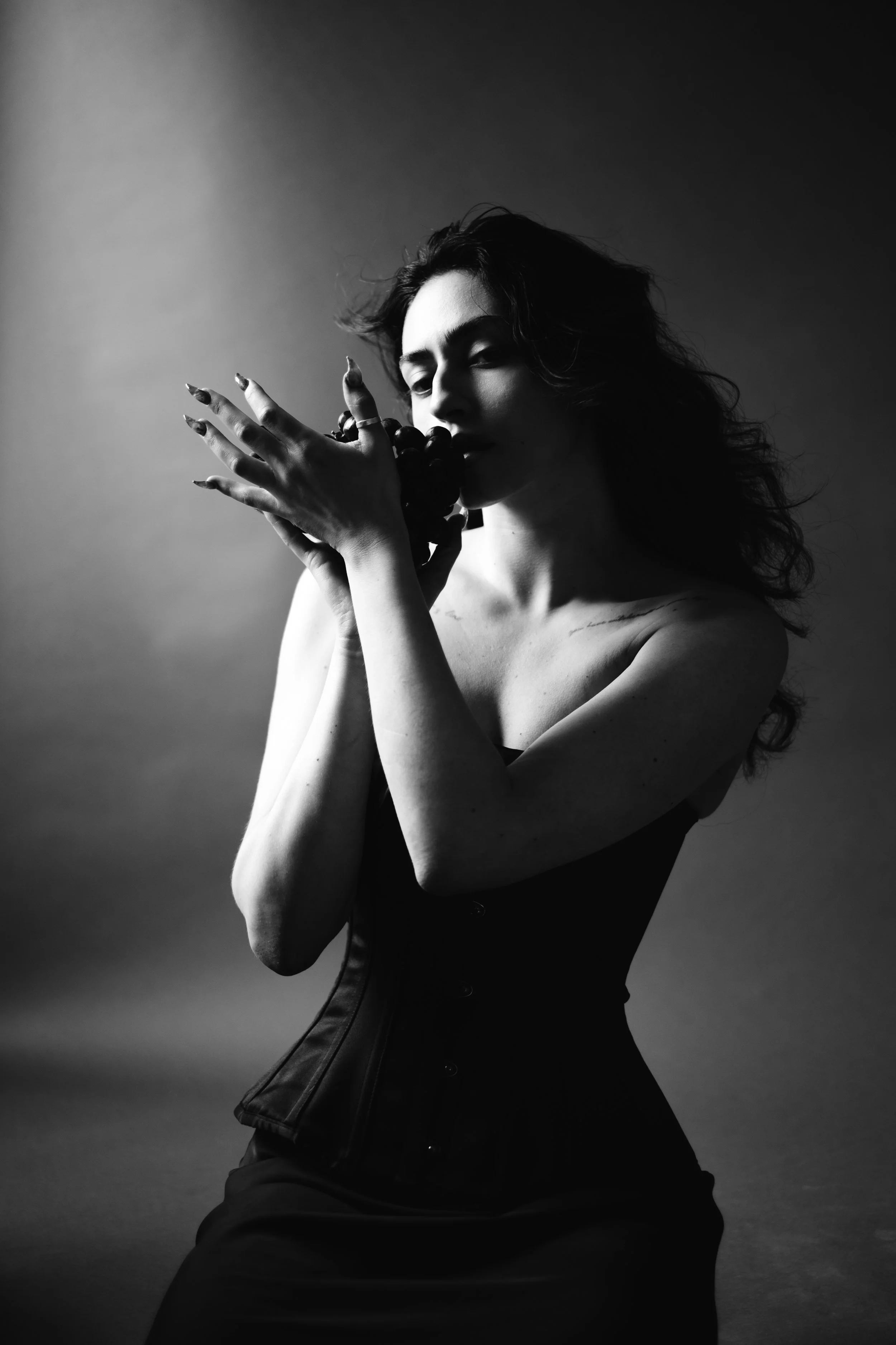 Black and white photo of a woman with dark curly hair, partly covering her face, posing with her eyes closed, holding grapes near her mouth, wearing a formal corset-style dress, against a plain gradient background.