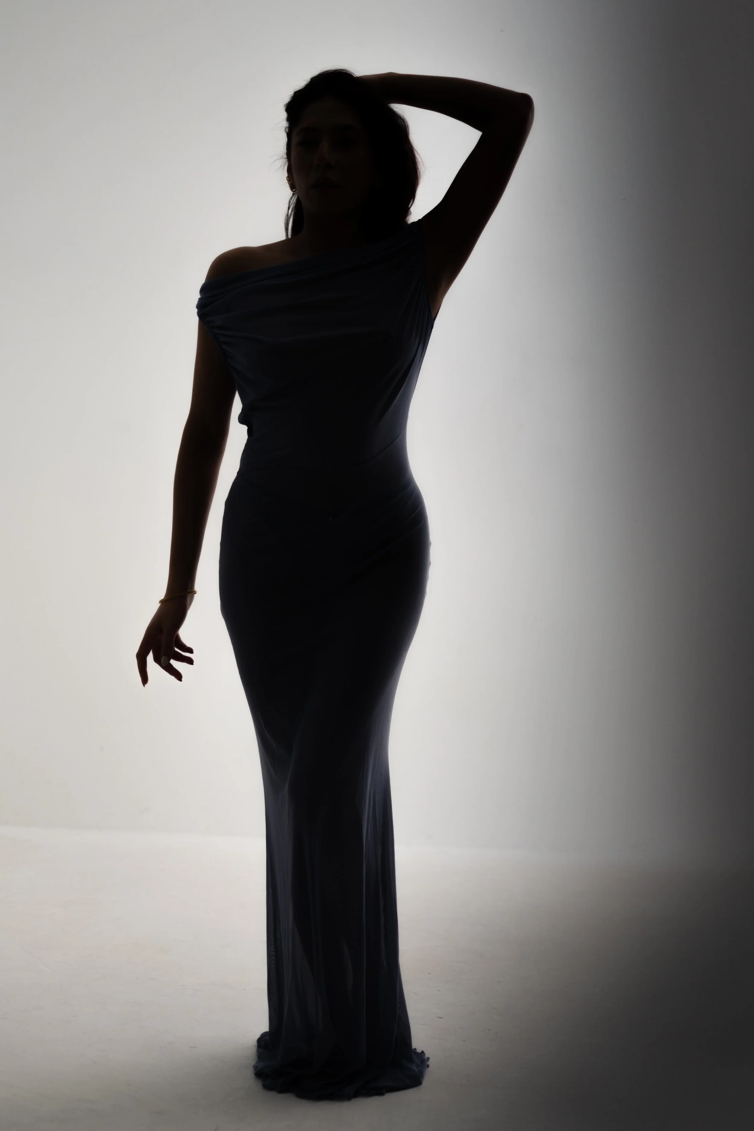 Silhouette of a woman standing with one hand behind her head, wearing a long, elegant dress in front of a plain, light background.
