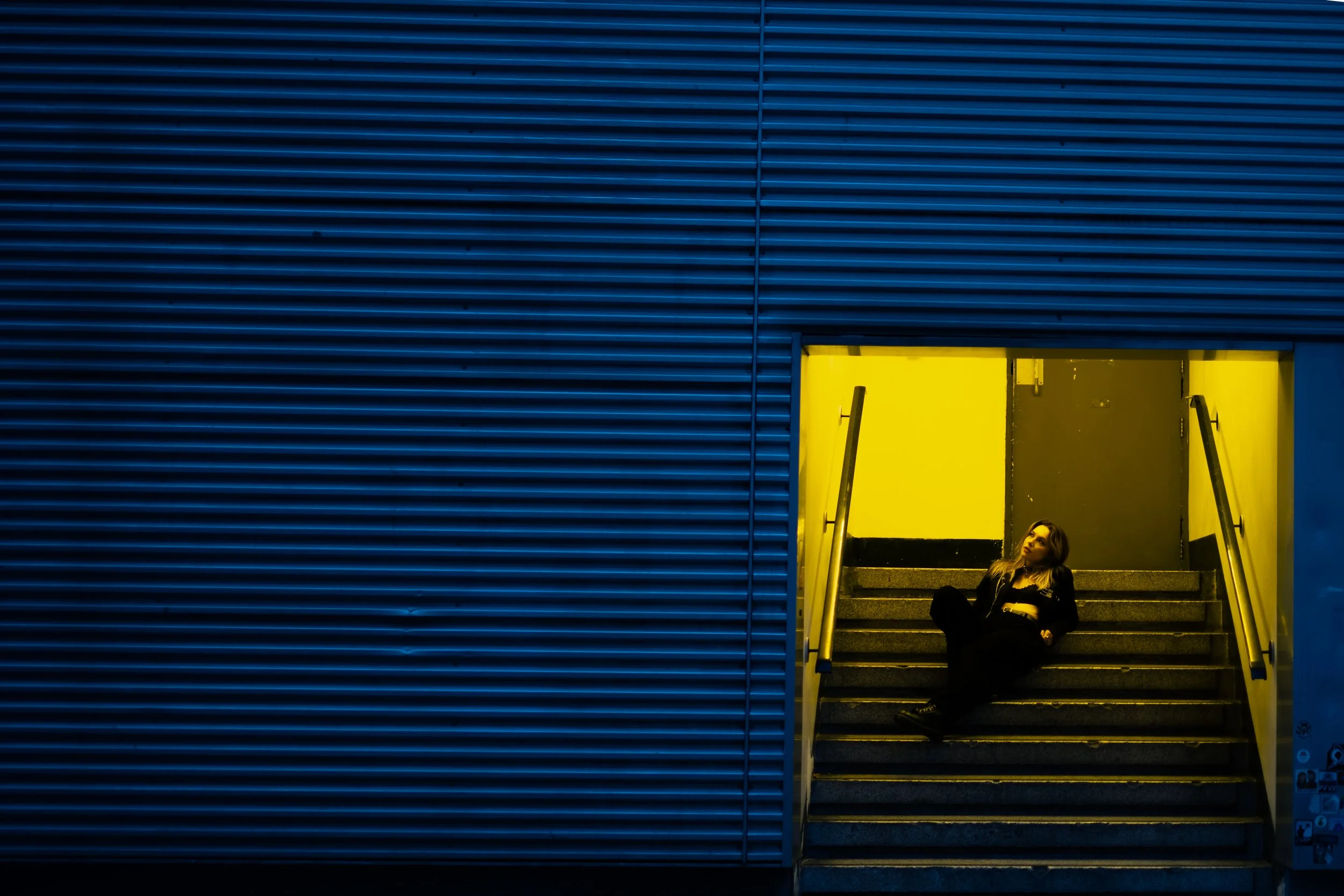 A young woman sitting on steps under a yellow-lit entrance against a dark blue wall with horizontal lines.