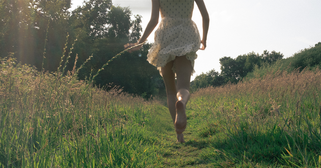 A person running through a grassy field during sunset, wearing a white dress with black polka dots.