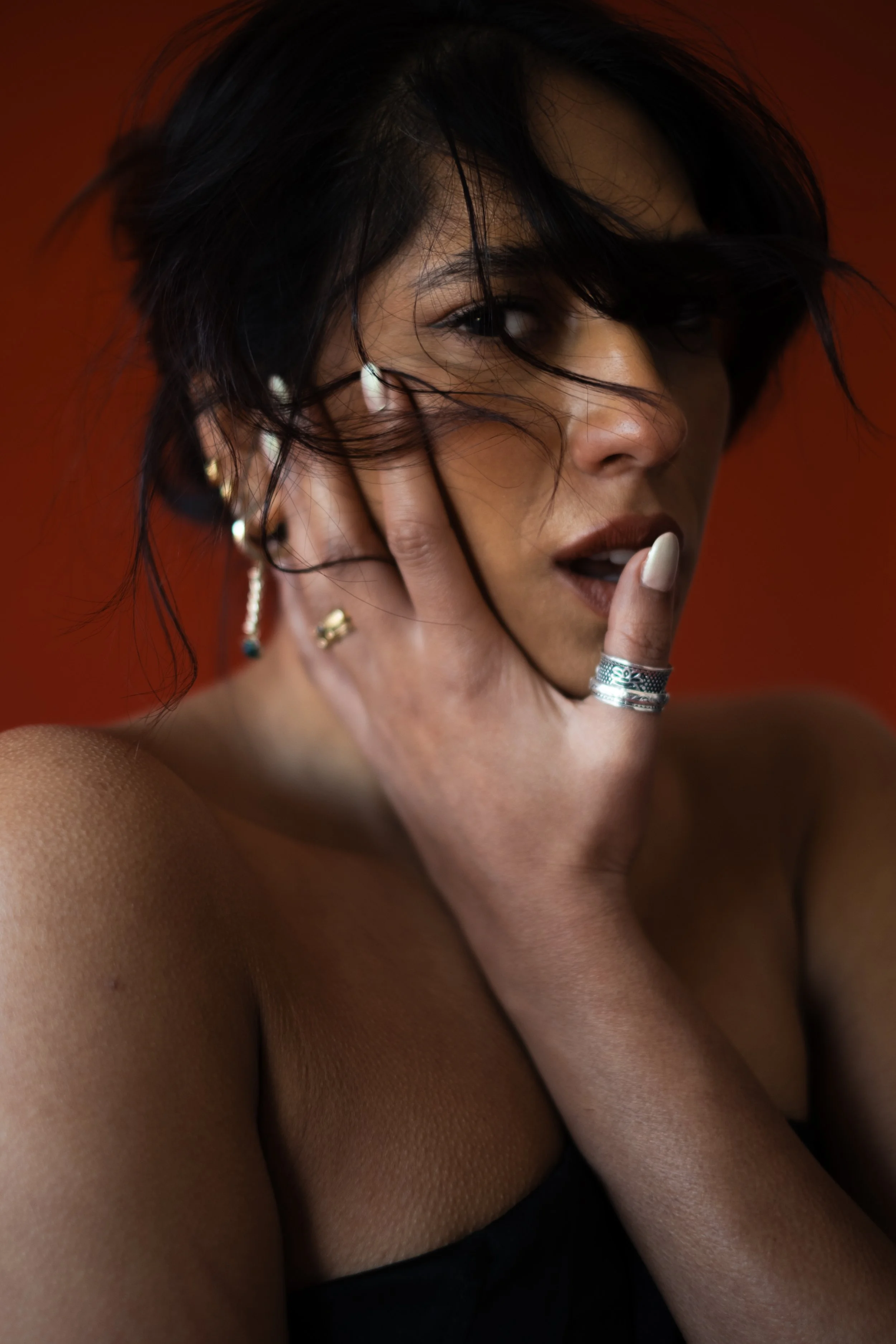 Woman with short dark hair and earrings, touching her face with her right hand, wearing rings, against a warm reddish-brown background.