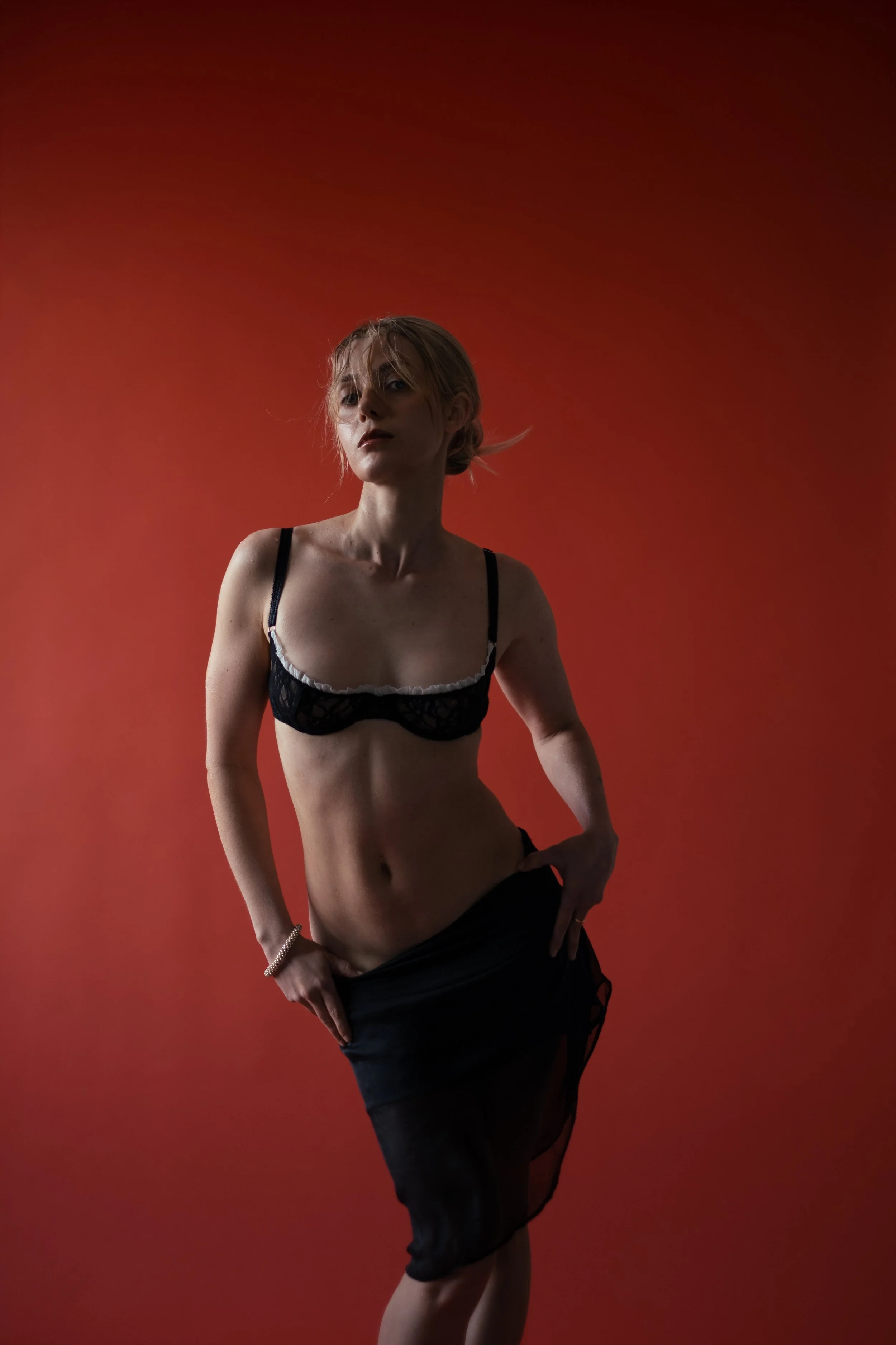 A woman with short blonde hair body-painted to look like she is wearing black lingerie against a red background.