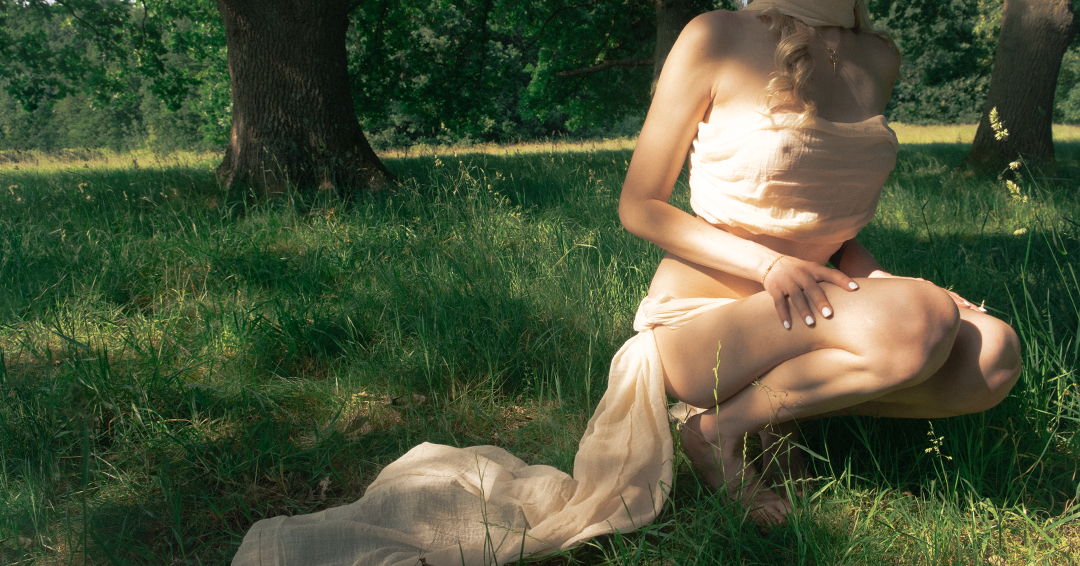 A woman kneeling in a grassy area with trees, wearing a beige dress, with her face not visible.