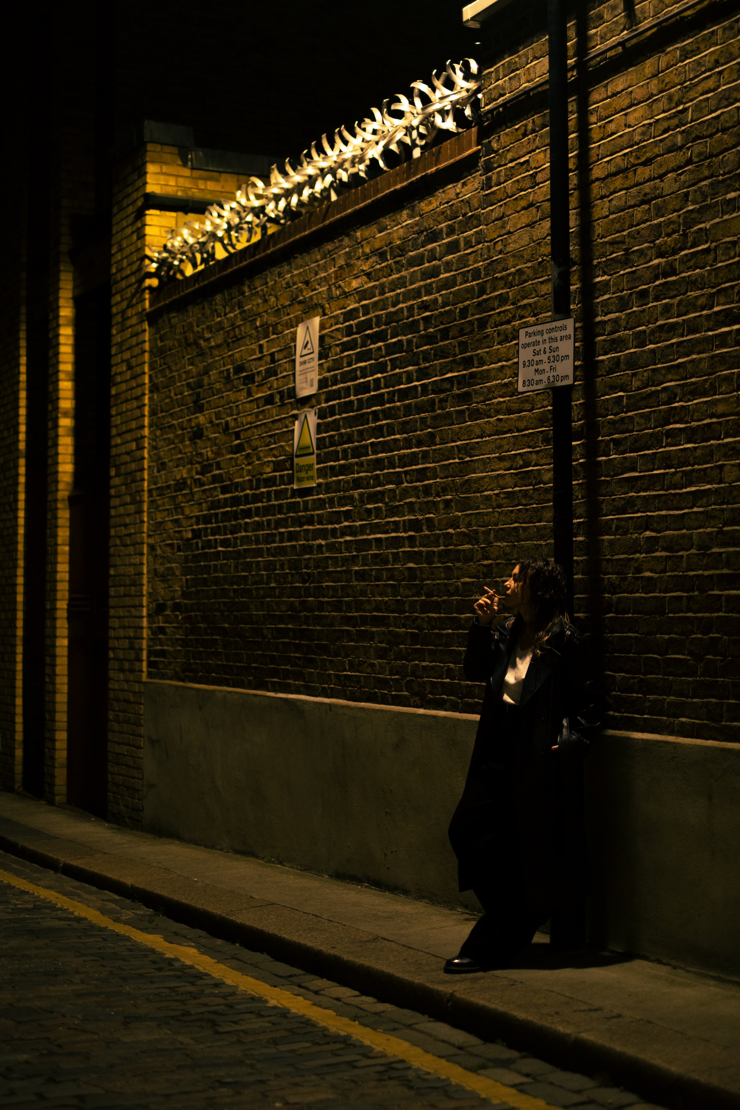 Woman leaning against a brick wall smoking at night, illuminated by a streetlight with signs on the wall.