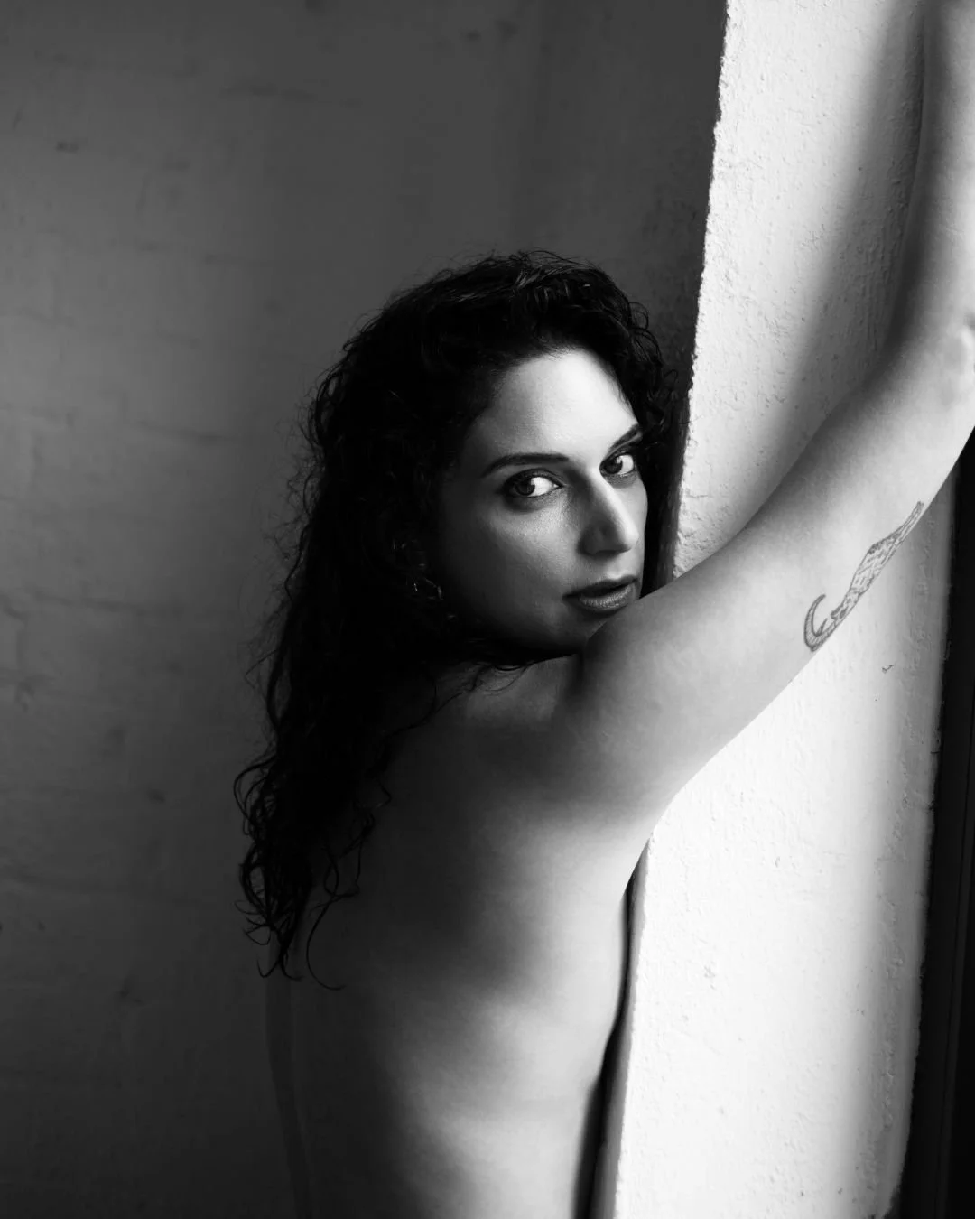 A black and white photo of a woman with curly hair, looking over her shoulder towards the camera, positioned near a wall.