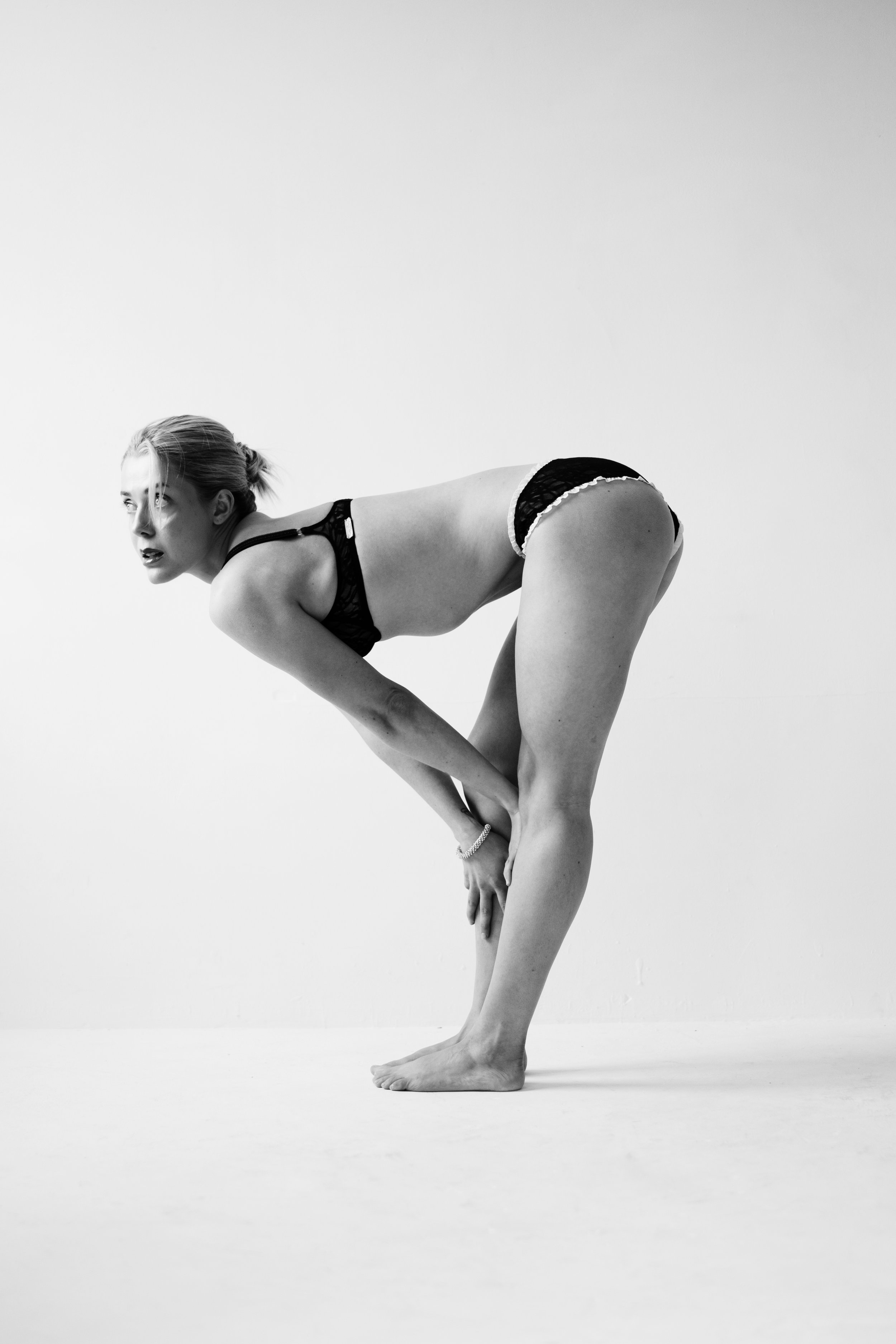A woman wearing black lingerie with lace and satin ruffle trim, bending forward with one hand on her ankle and the other on her knee, against a plain white background.
