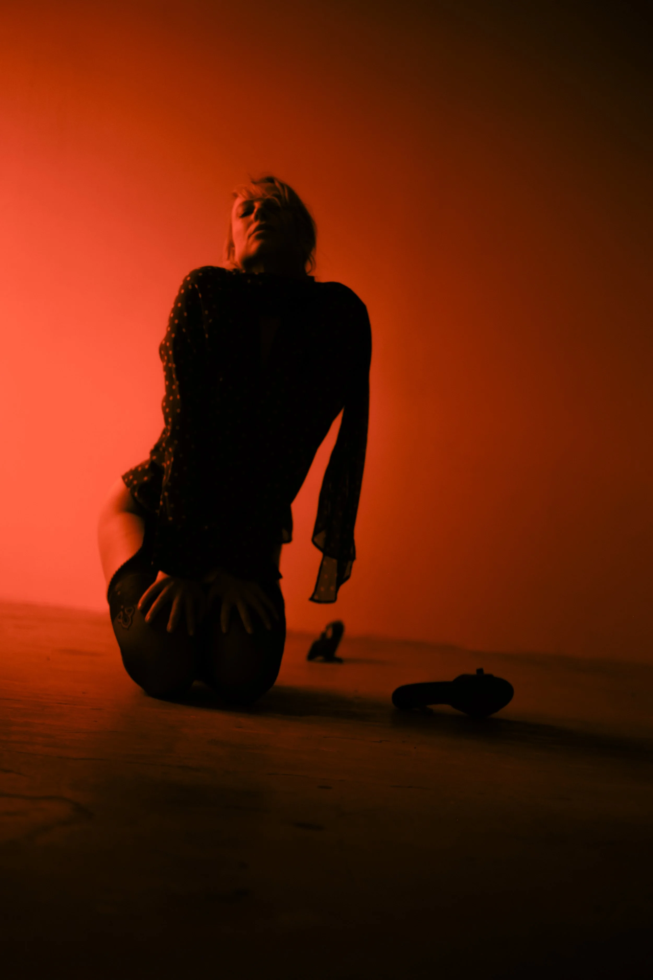 A person kneeling on the ground with their head tilted back, illuminated by warm reddish-orange lighting, in a setting that appears to be a beach or desert. The person wears a polka dot top and has tattoos on their thigh, with objects placed on the g
