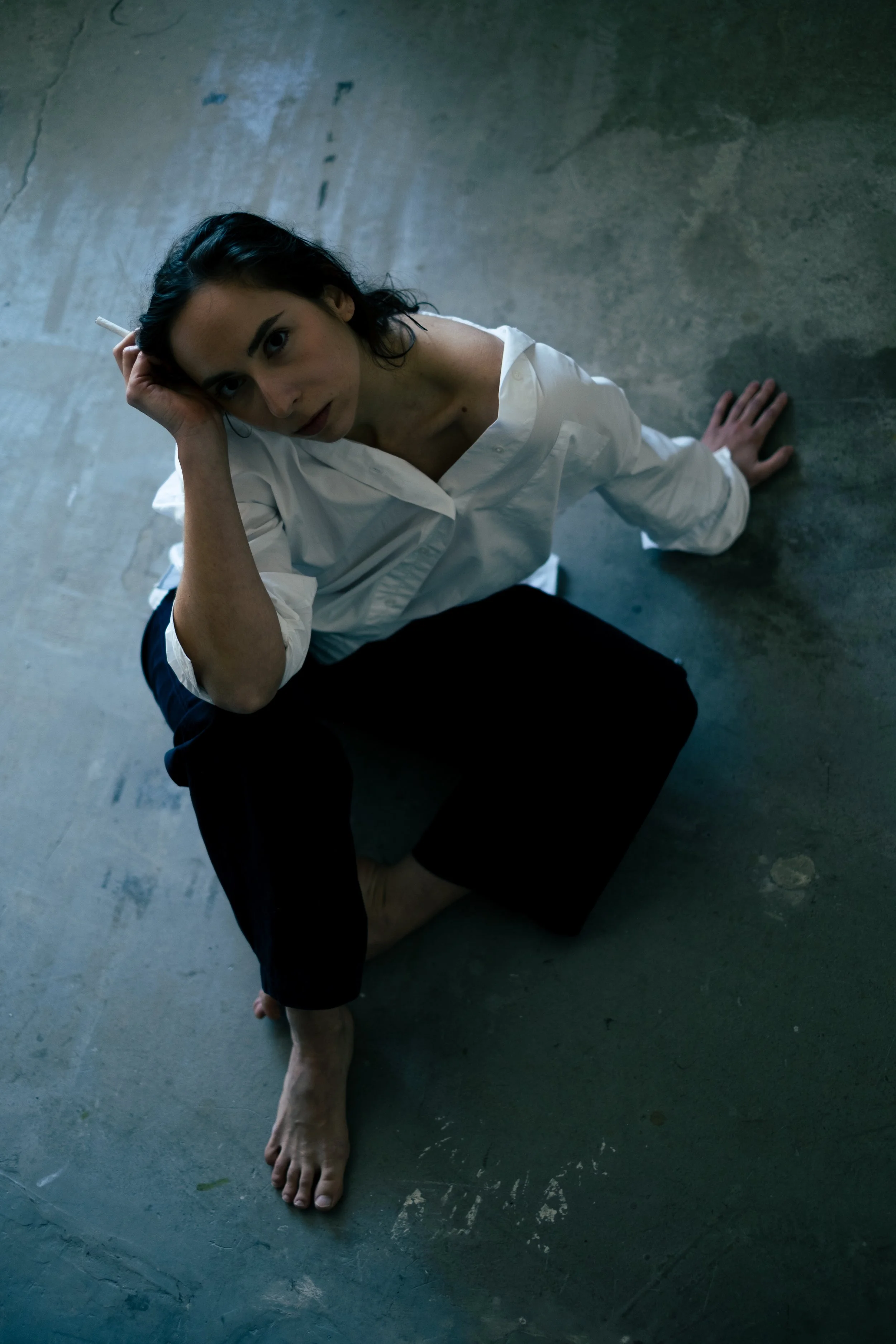 A woman with dark hair and dark eyes, wearing a white shirt and black pants, sits on a concrete floor with her legs folded beneath her. She is looking up at the camera, with one hand resting on her head and the other on the ground.