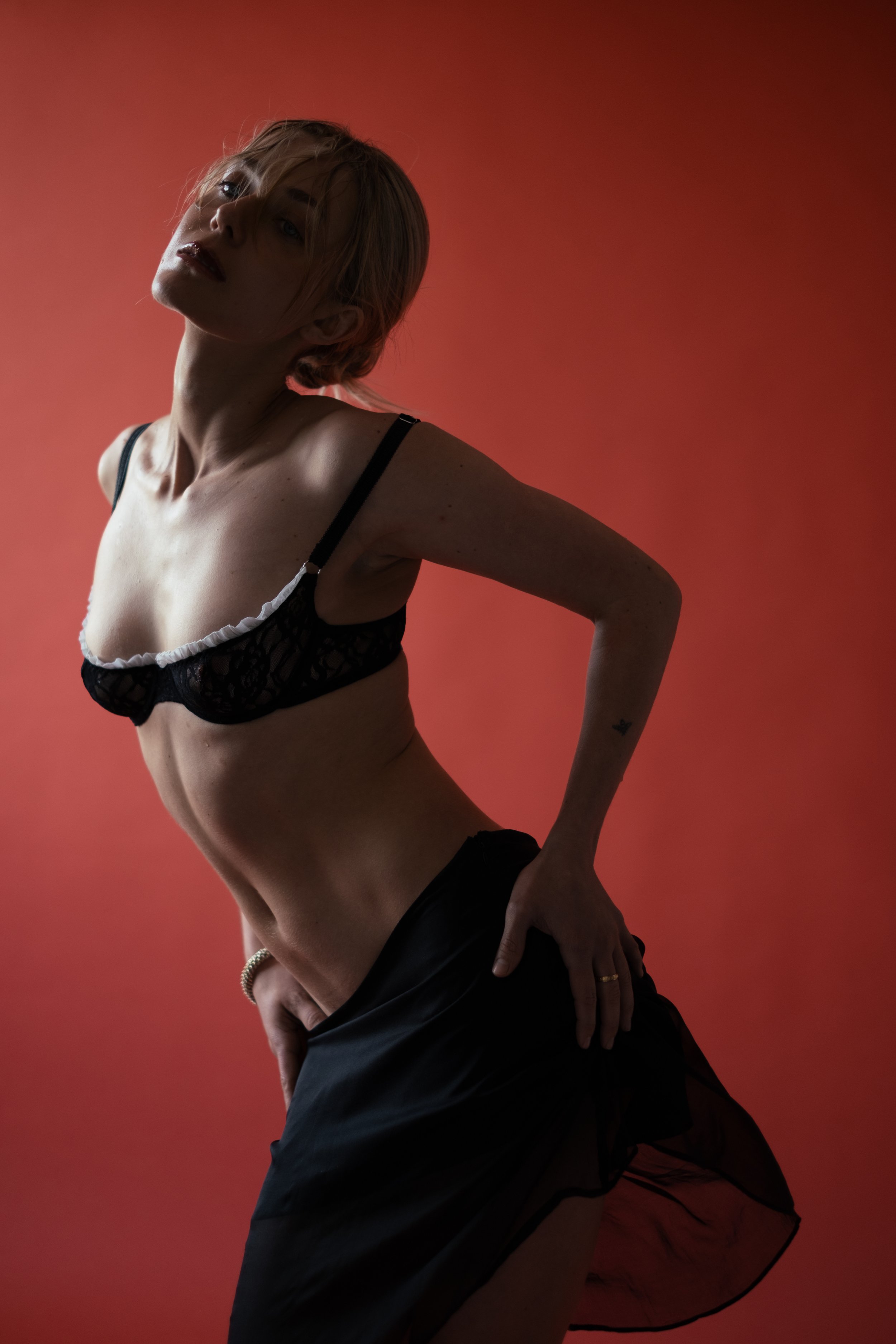 A woman with short blonde hair posing against a red background. She is wearing black lingerie with lace details and a black sheer skirt. She has a confident expression and is looking slightly upward with her left arm bent and hand on her hip.