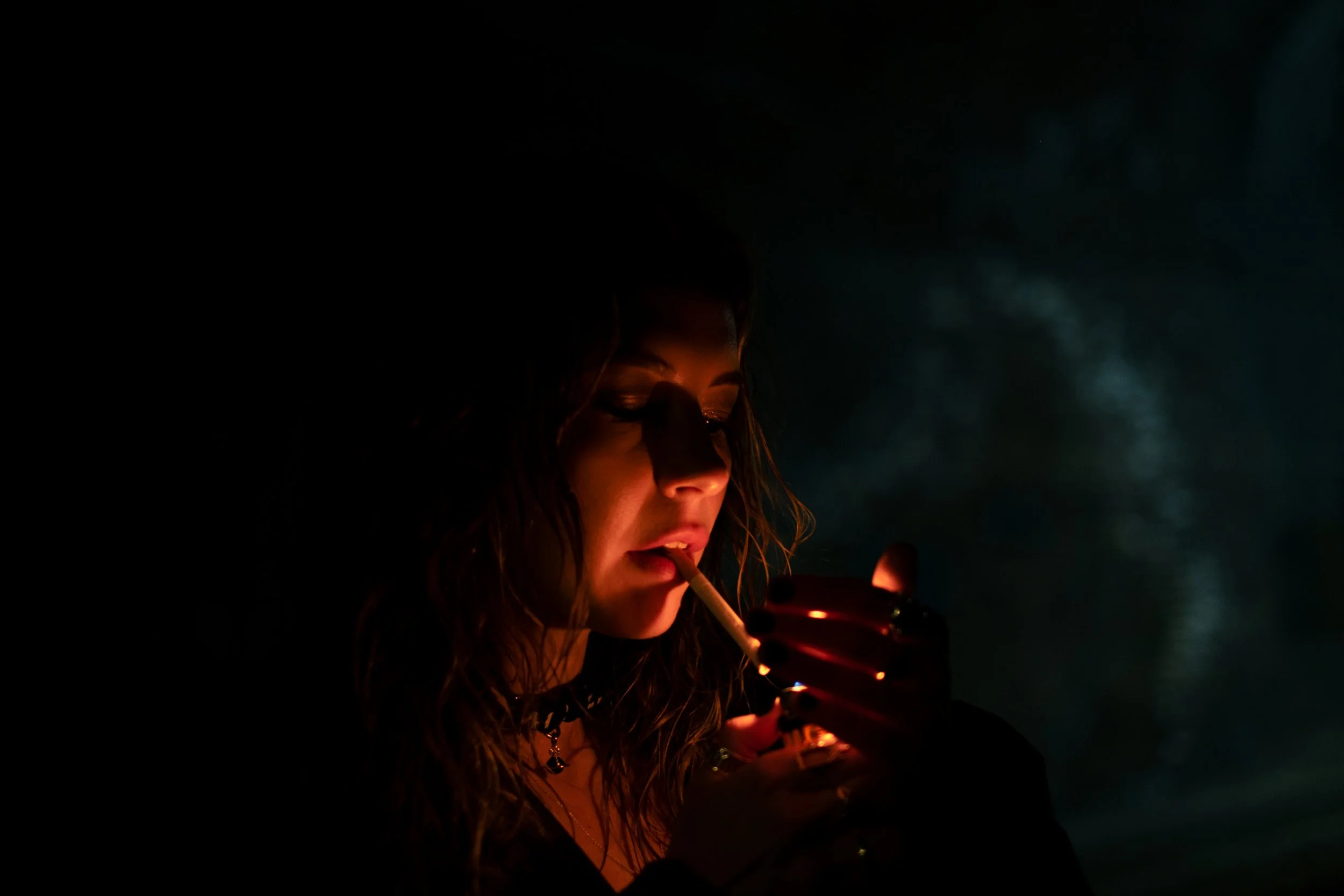A woman with curly hair in a dark setting, illuminated by the glow of a lighter or match, lighting a cigarette.