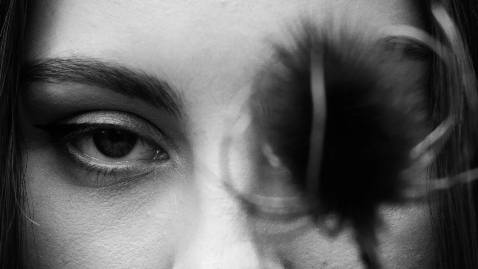Close-up black and white photo of a person's face with one eye visible and a large dark feather or leaf obscuring part of their face.