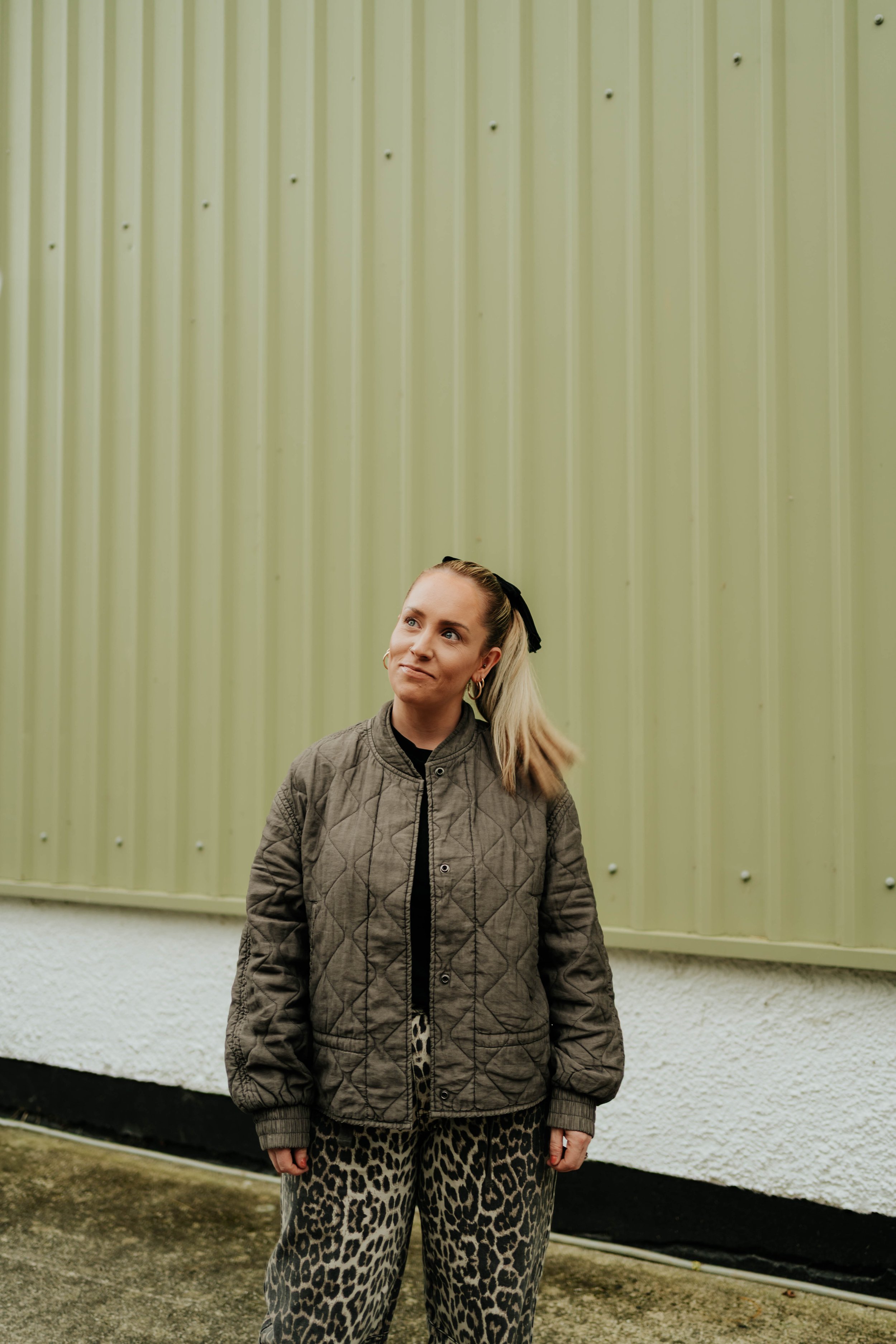 Woman standing outdoors in front of a green metal wall, wearing a quilted brown jacket and leopard print pants, with her hair tied back and wondering expression.