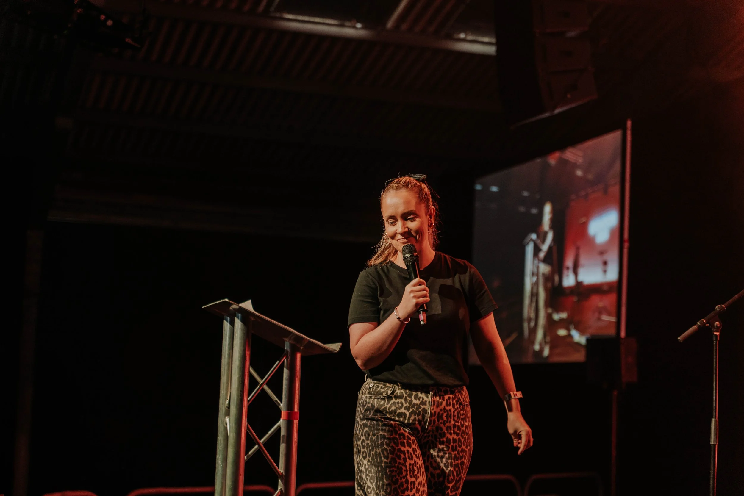 A young woman with brown hair in a ponytail is standing on stage, holding a microphone, and smiling. She is wearing a black t-shirt and leopard print pants, with a watch on her left wrist. There is a podium to her right and a large screen behind her displaying her image.