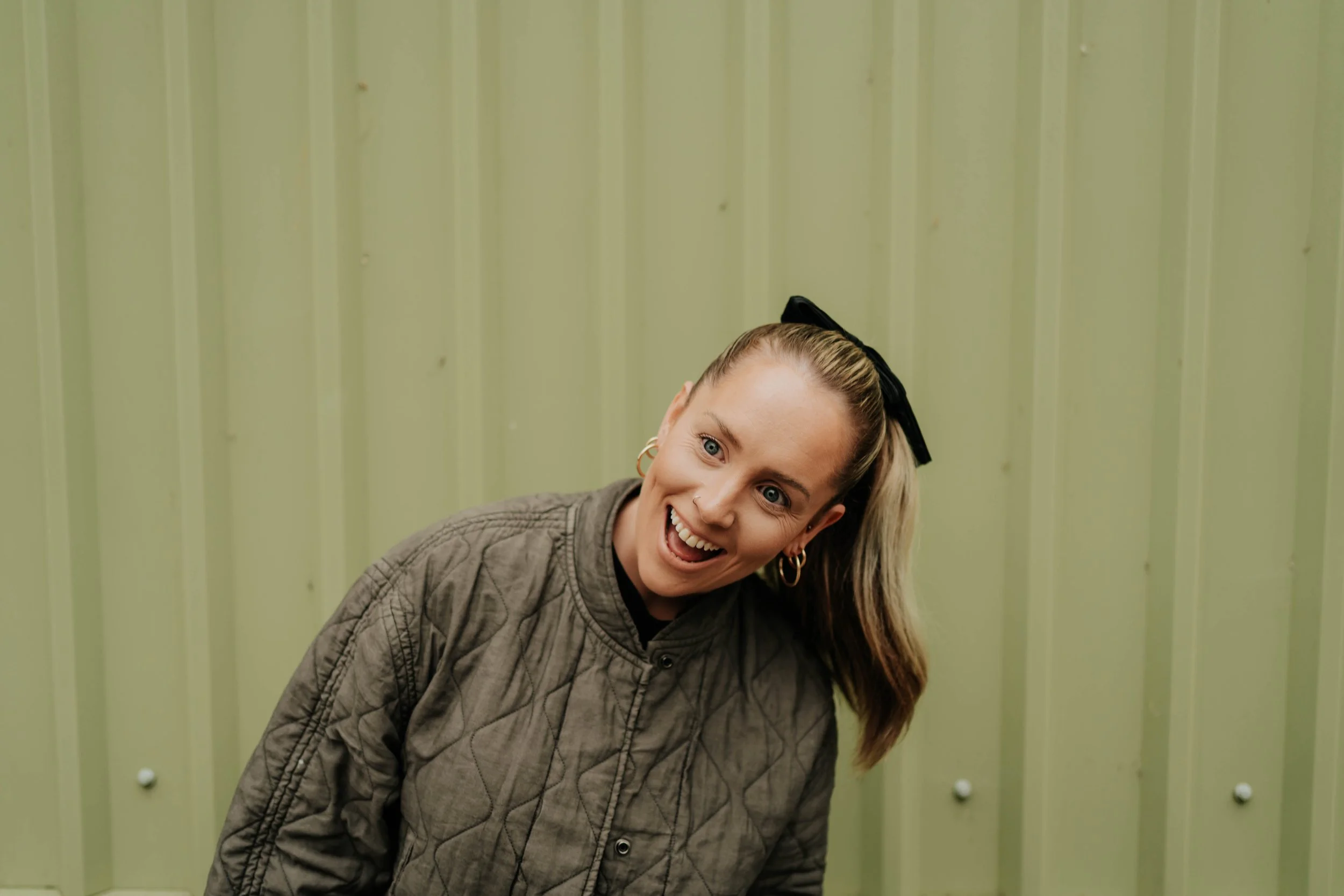 A woman with blonde hair styled in a high ponytail secured with a black bow, smiling excitedly, wearing a quilted gray jacket and hoop earrings, standing in front of a light green metal wall.