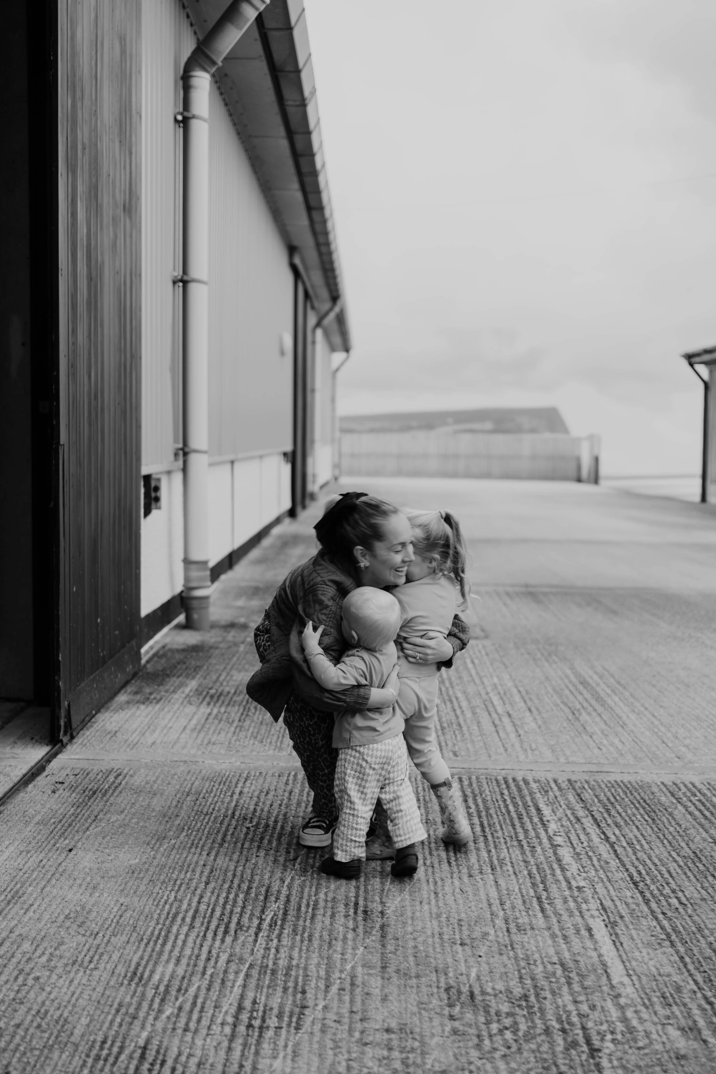 A woman and two children hugging and smiling outside a barn-like building on a concrete surface, in black and white.