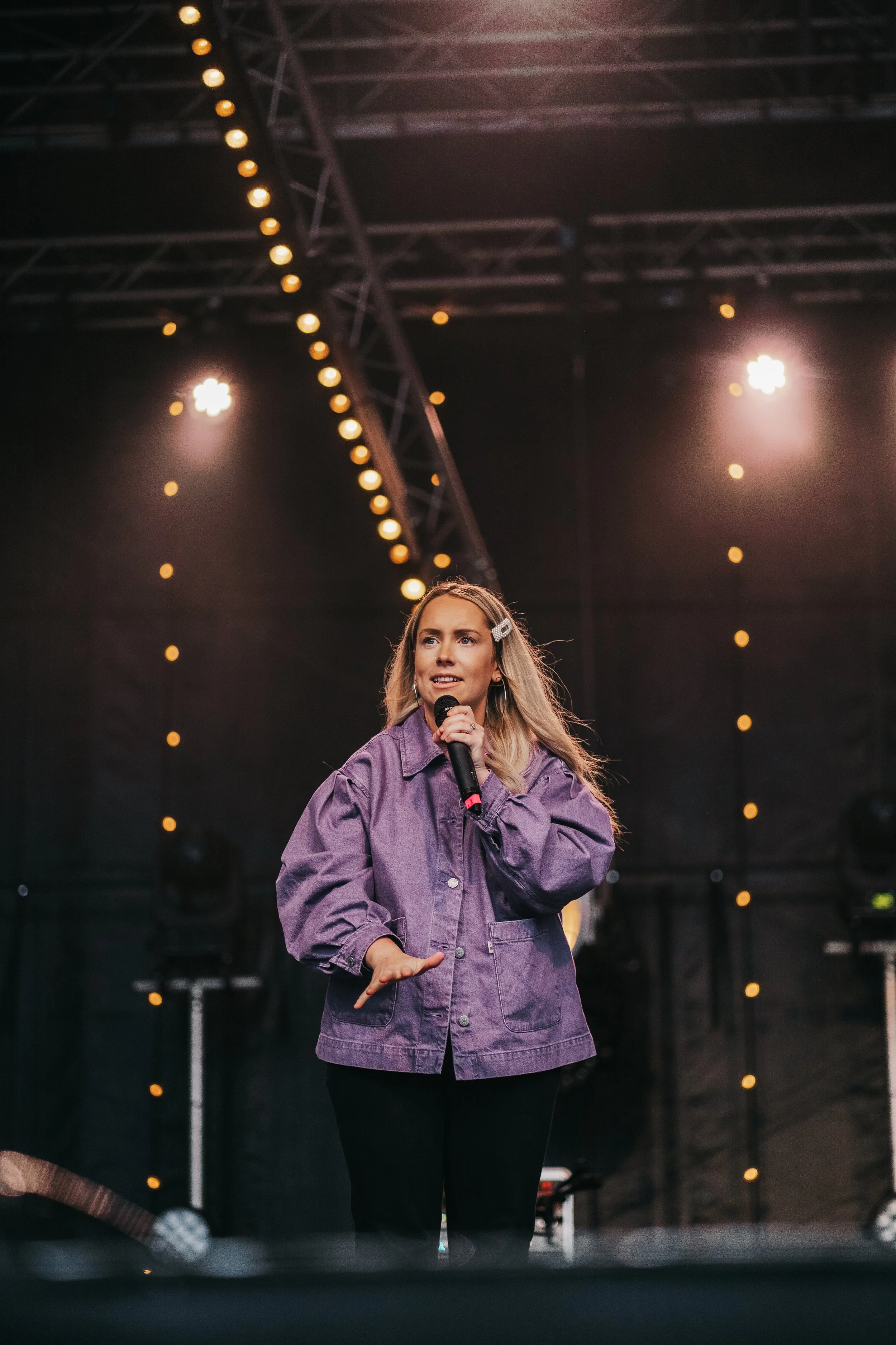 A young woman with long blonde hair, wearing a purple oversized denim jacket and black pants, singing into a microphone on stage with a dark background and string lights overhead.