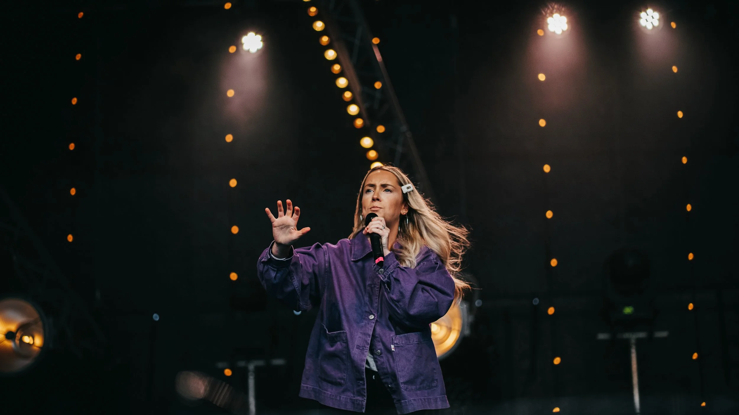 A woman singing into a microphone on stage with bright spotlights and decorative lights behind her.