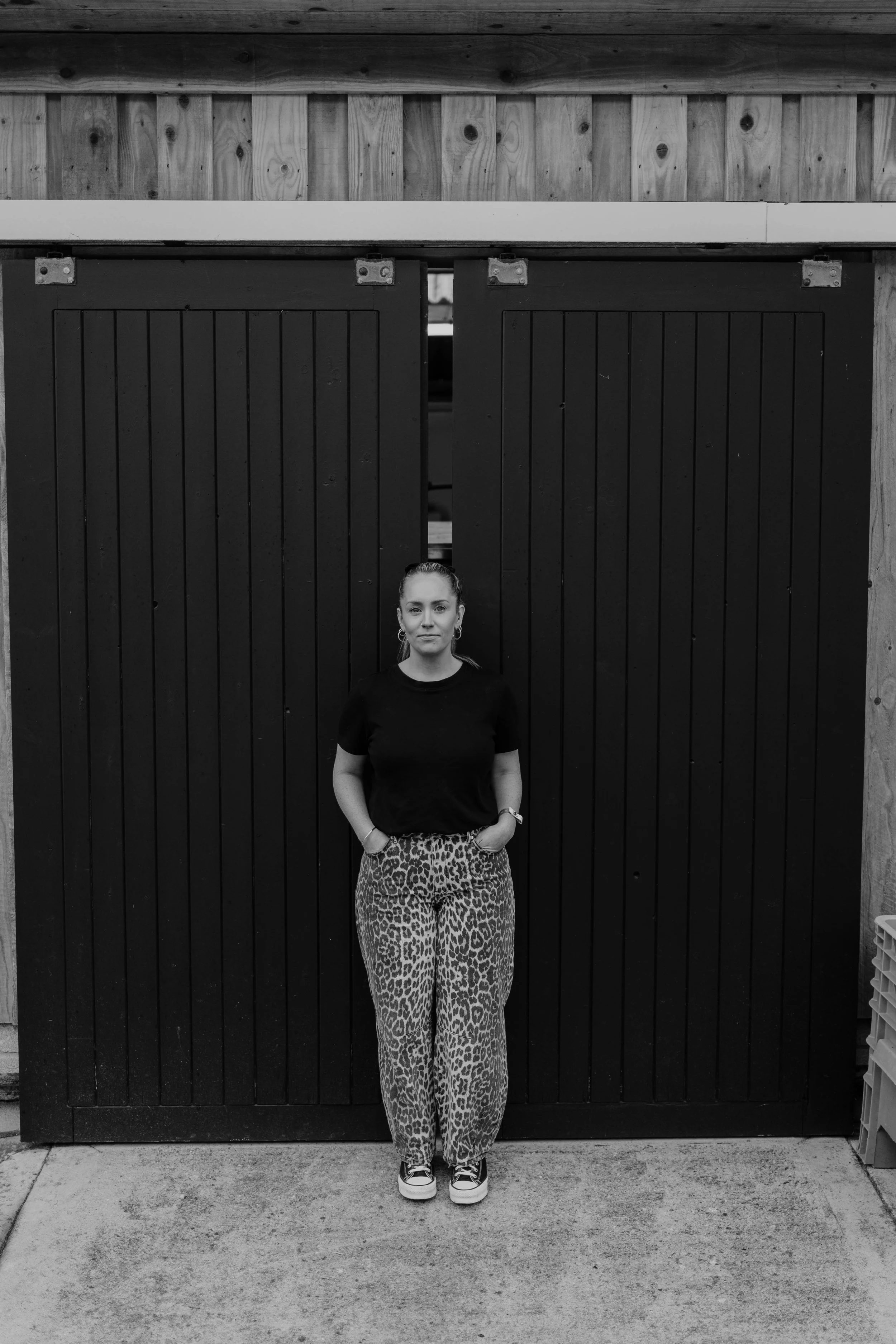 A woman standing in front of a black barn door with wooden framing, wearing a black T-shirt, leopard print pants, and sneakers, with her hands in her pockets.
