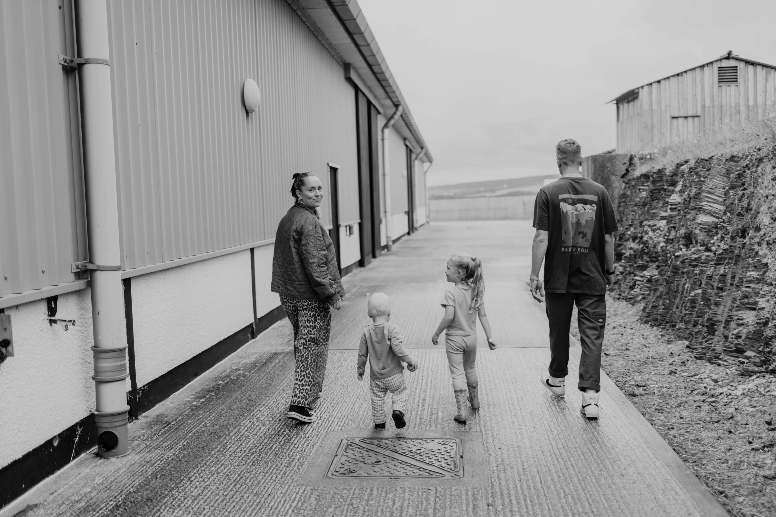 A black and white photo of a family walking outdoors on a paved path beside a metal building and a gravel hill. The family includes a woman, a man, and two young children. The woman looks back and smiles at the camera while the man walks ahead. The children walk between the adults, one holding the other's hand.