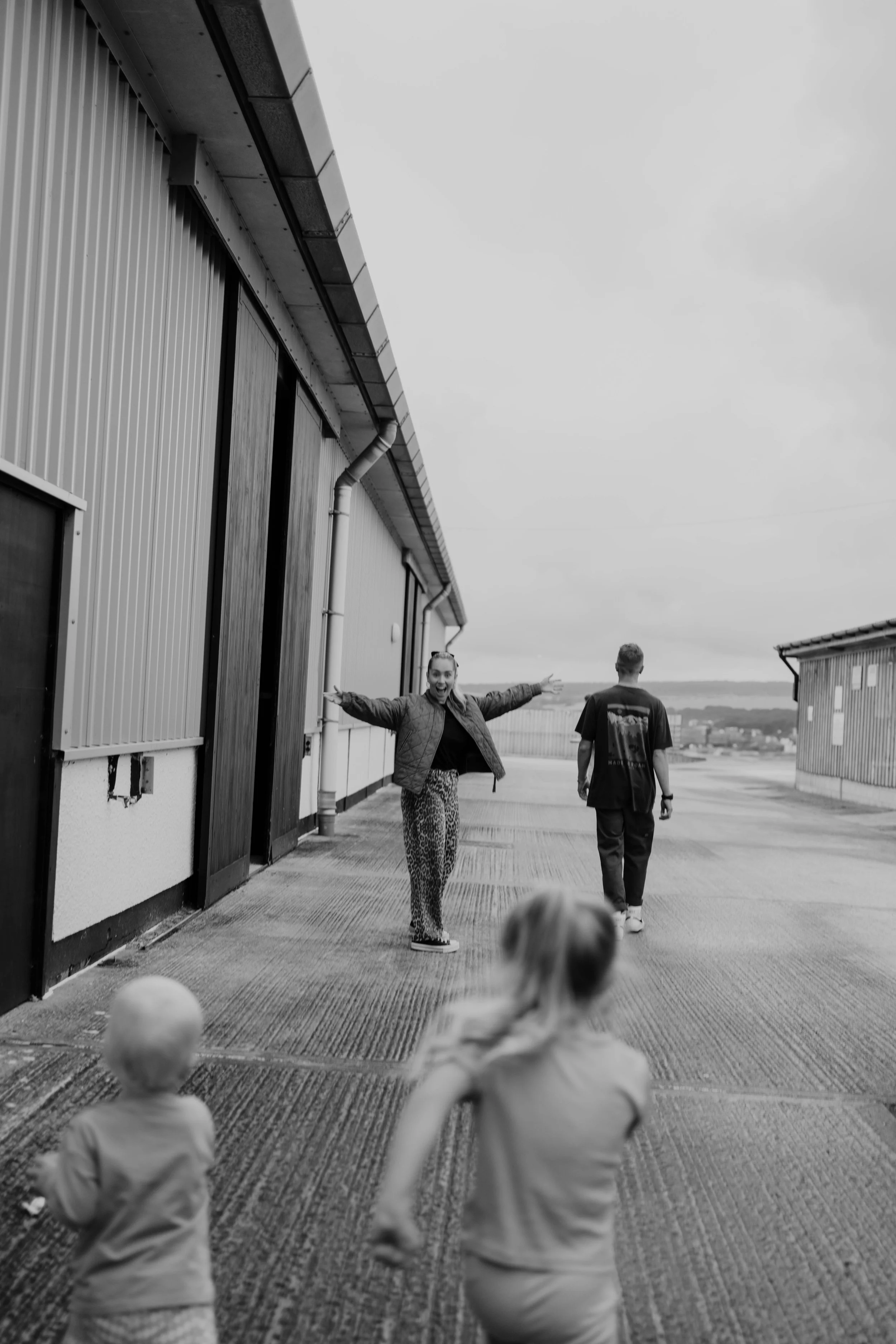 A woman happily engaging with children outside on a concrete surface next to a large industrial building with metal siding, while a man walks away in the background.