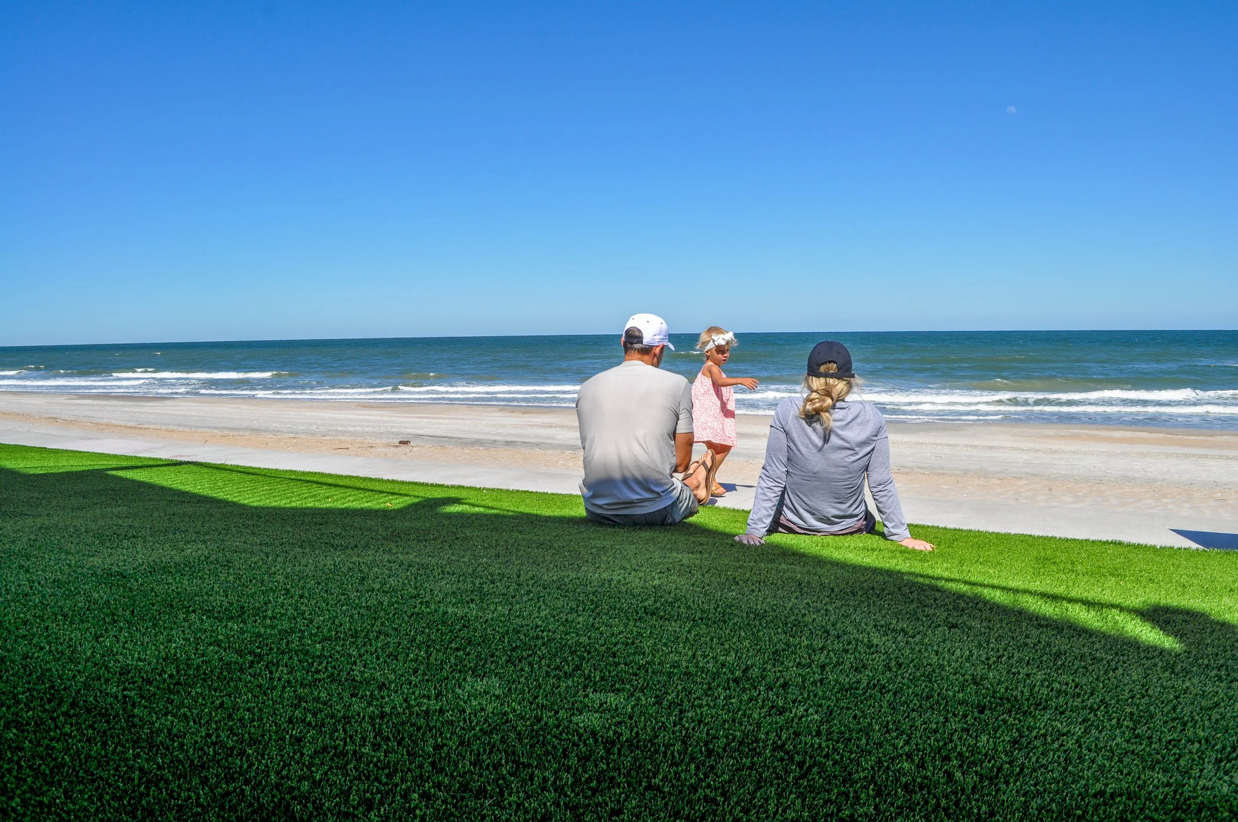 A family of three sitting on artificial grass near a beach, with a man, woman, and small girl playing near the shoreline, under a clear blue sky.
