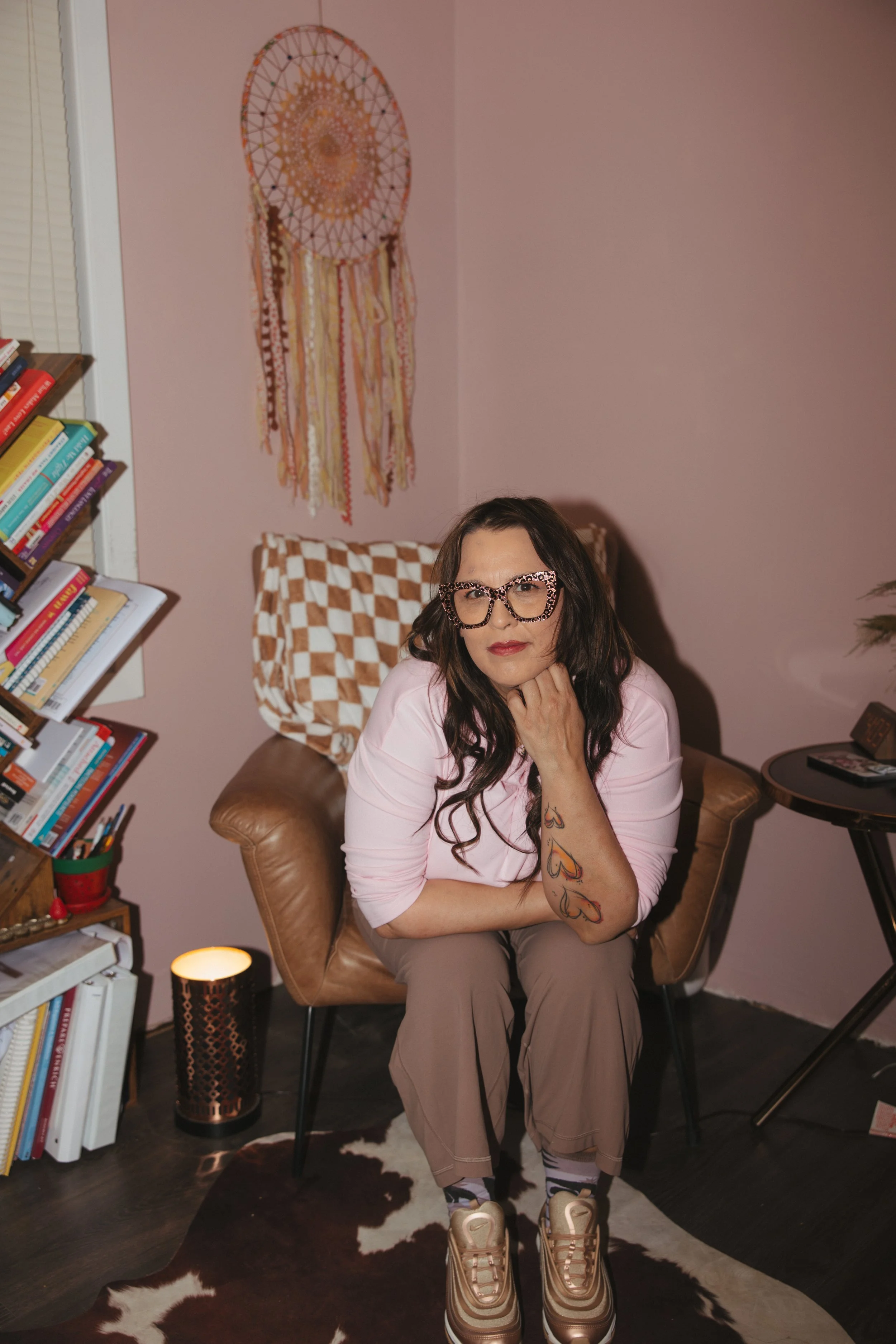 A unique woman coach and therapist who is quirky with a wild streak, sitting in a cozy room with a pink wall, a bookshelf filled with colorful books, and artistic decor.