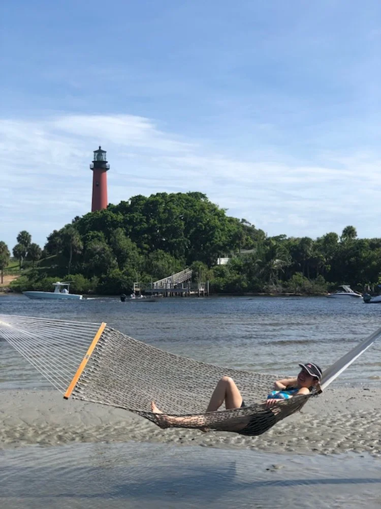 Maria Perdomo-Torres, Cuban therapist, relaxing in a hammock by the beach in Florida, with a lighthouse in the background. Symbolizing rest, balance, and the calm therapy can bring.