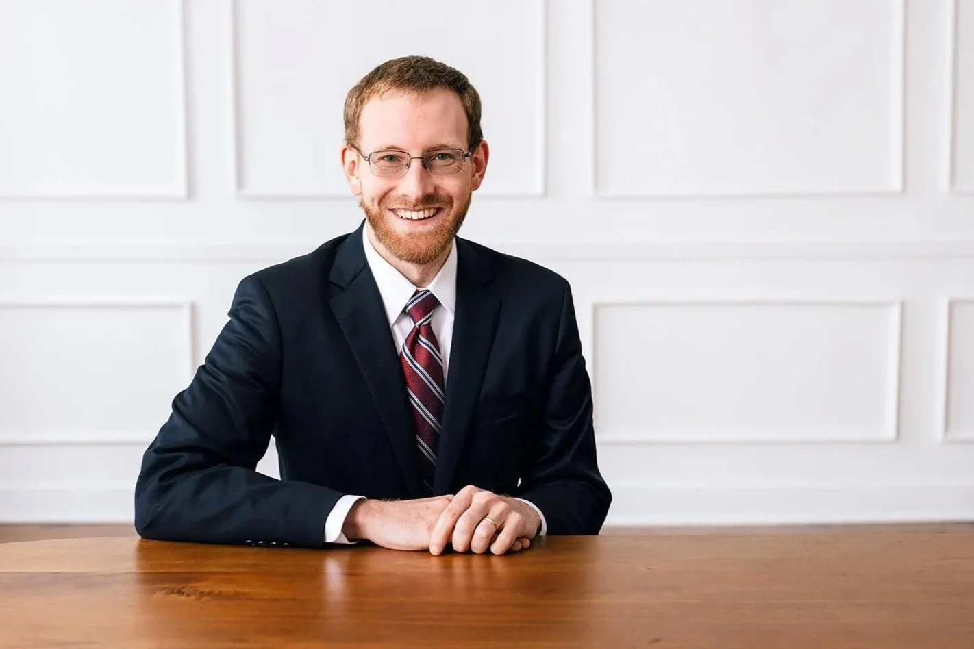 Brenden Ponder man with short brown hair, glasses, and a beard sitting at a wooden table, smiling at the camera, wearing a dark suit, white shirt, and a striped tie, in a white-paneled room.