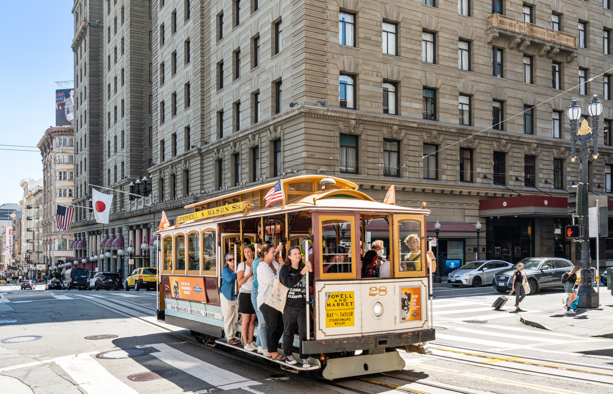 San Francisco cable car with passengers crossing a city street, surrounded by tall historic buildings, American and Japanese flags flying overhead, and pedestrians walking.