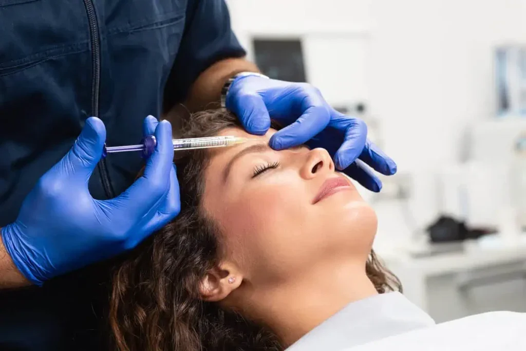 A woman receiving a cosmetic injection in her forehead from a healthcare professional wearing blue gloves.