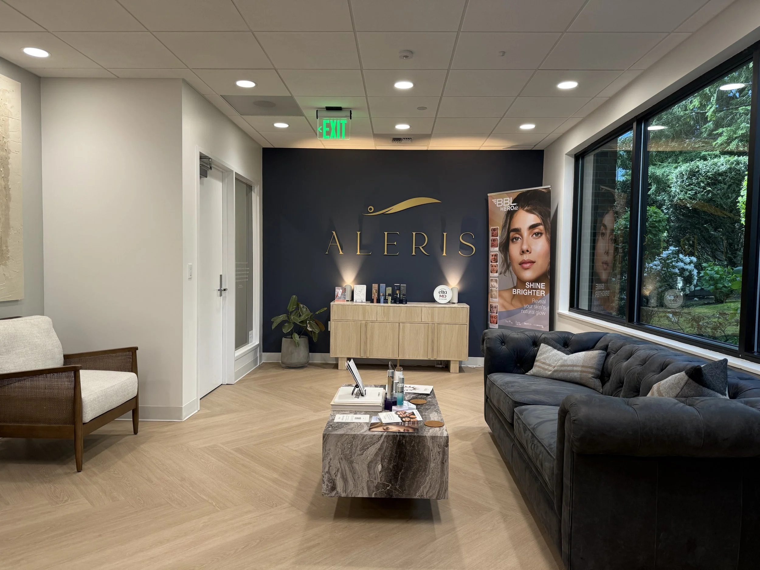 Interior view of a waiting or reception area with chairs, a coffee table, a plant, and a counter with beauty products. There is a large window with trees outside and a sign with the brand name 'Aleris' on the wall.