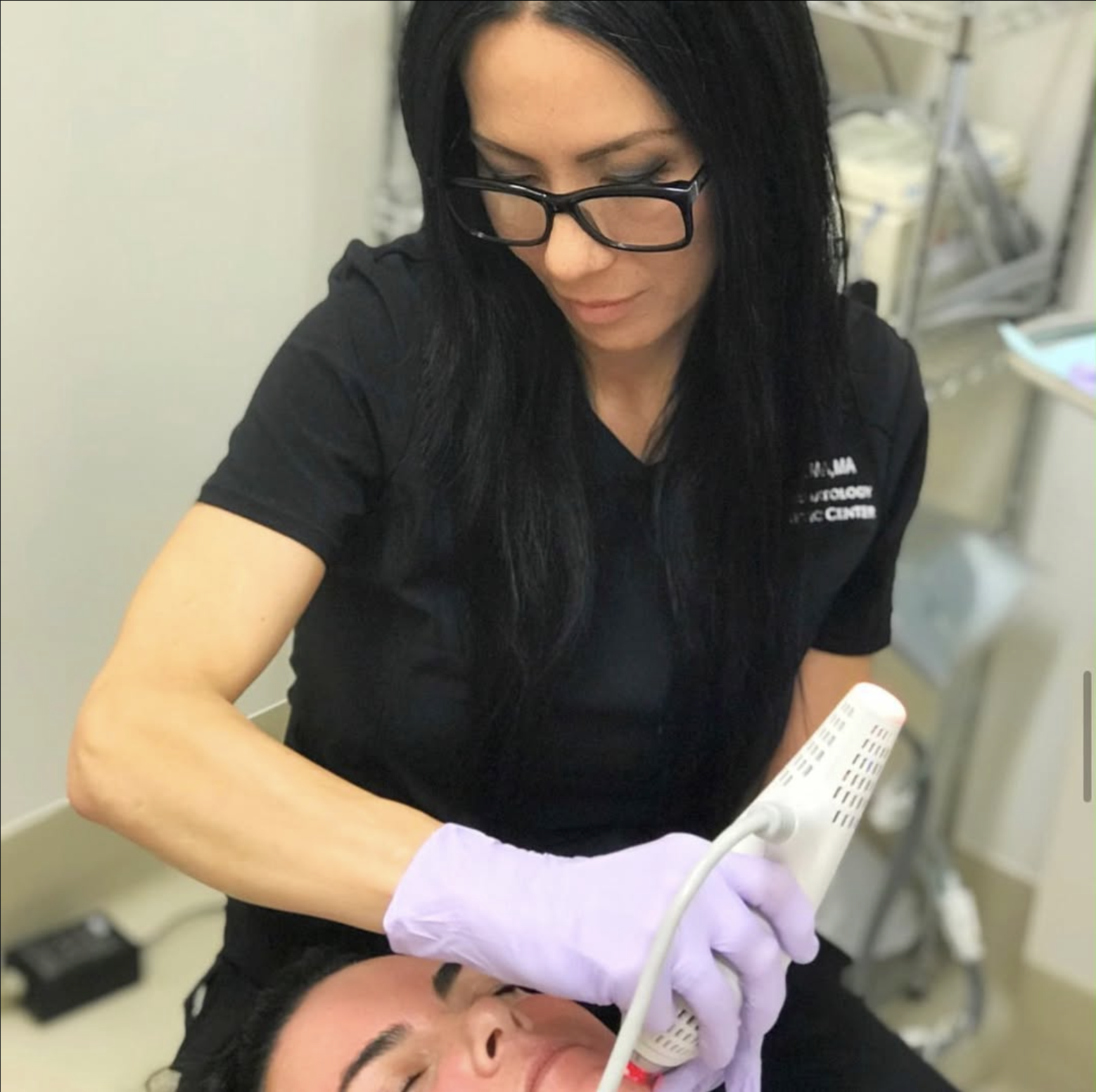 A female healthcare professional, wearing glasses and purple gloves, performs a procedure on a patient using a medical device in a clinical setting.