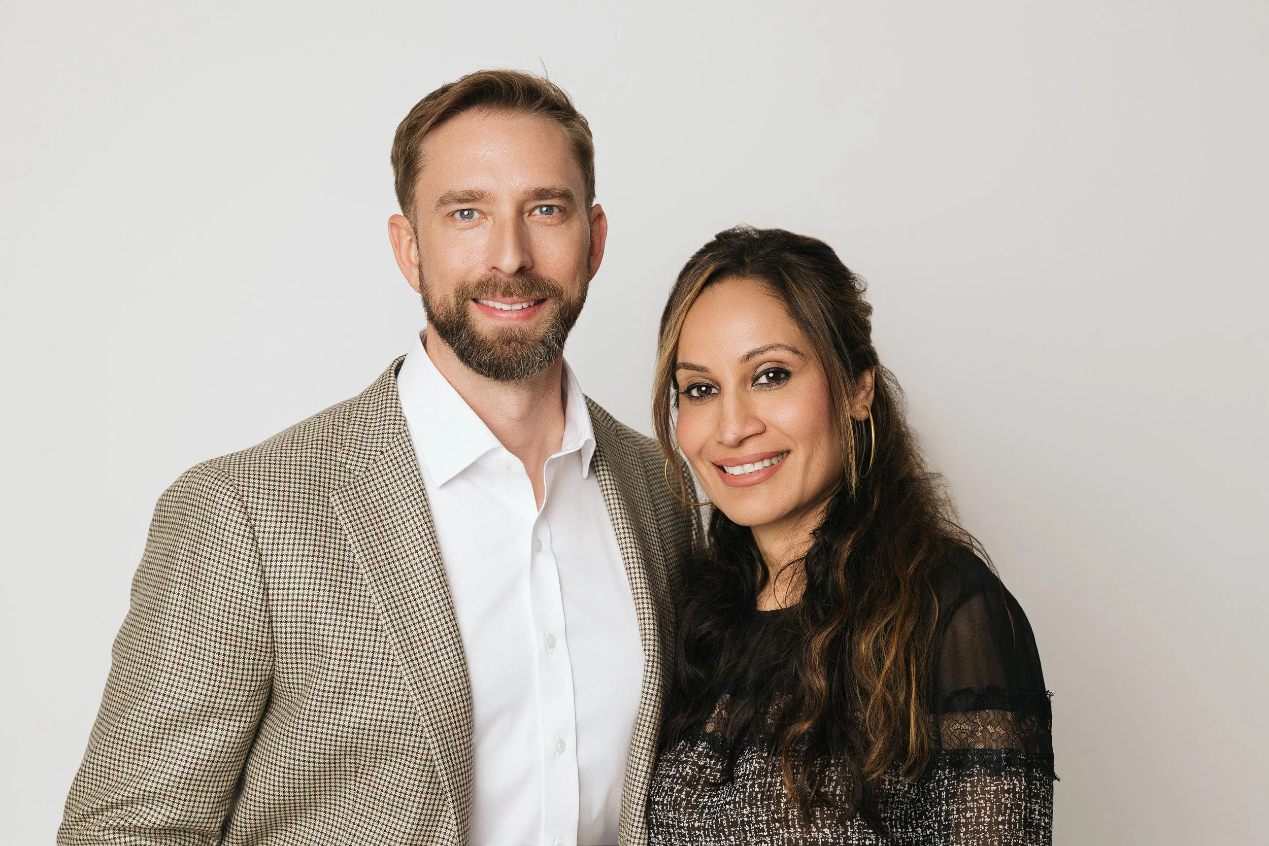 A man and woman smiling, standing side by side against a plain light gray background.