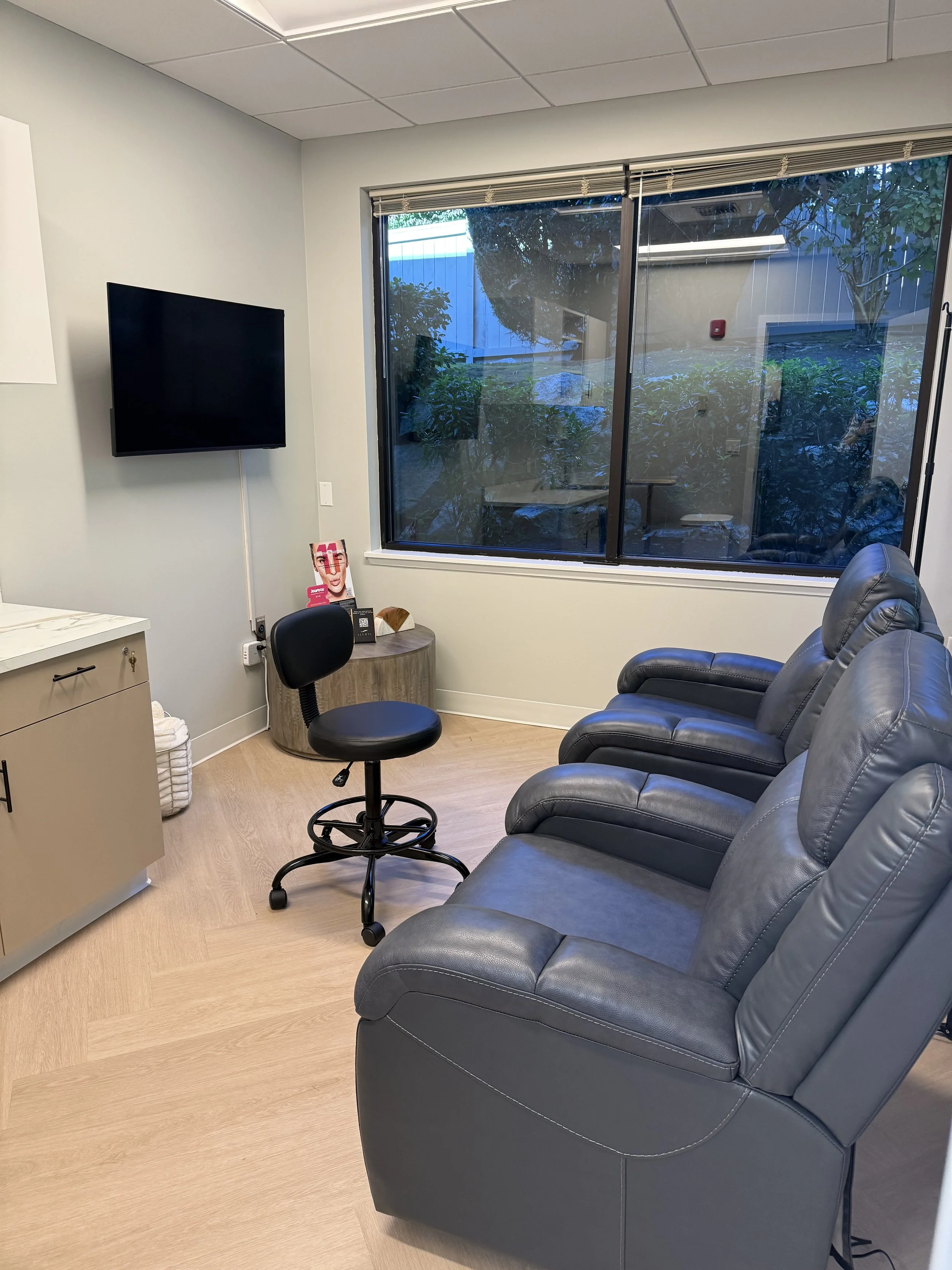 An office waiting room with three dark gray leather chairs, a black adjustable chair, a small wooden table with a magazine and a tissue box, a wall-mounted flat-screen TV, large window with outdoor plants visible, and light-colored wooden flooring.