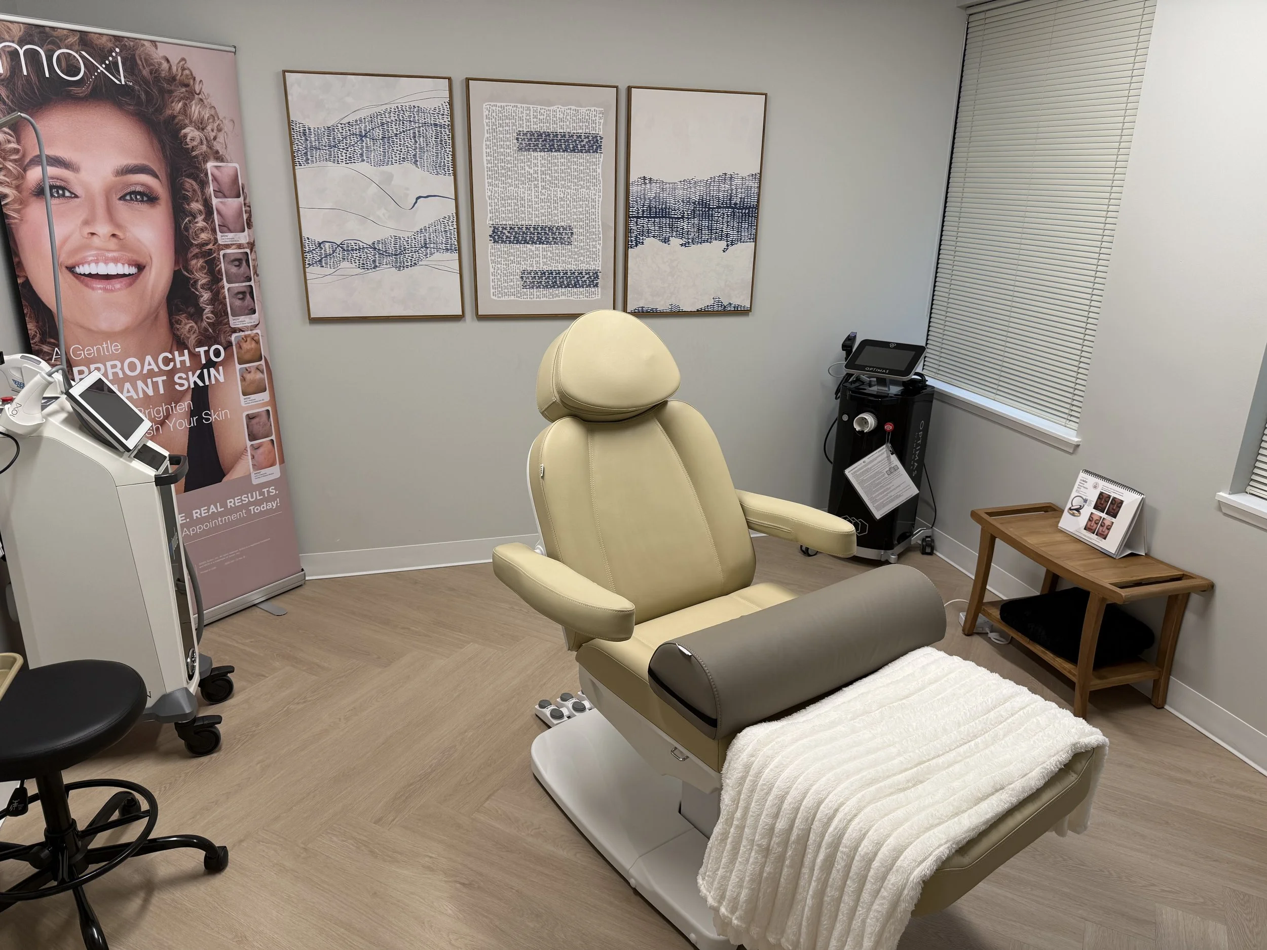 A medical examination room with a beige examination chair covered with a white blanket, a black stool, a medical device with a screen, a small wooden table with brochures, and three abstract framed artworks on the wall. A large poster of a smiling woman with curly hair promoting skin treatment is partially visible on the left side of the image.