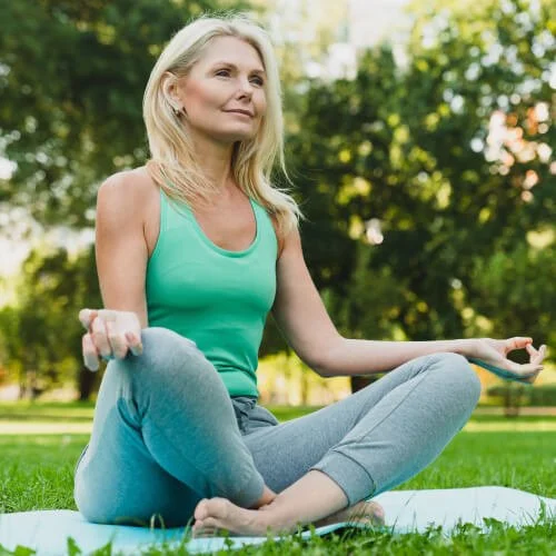 Woman practicing yoga outdoors in a park, sitting cross-legged on a mat, meditating with eyes open, surrounded by green trees.