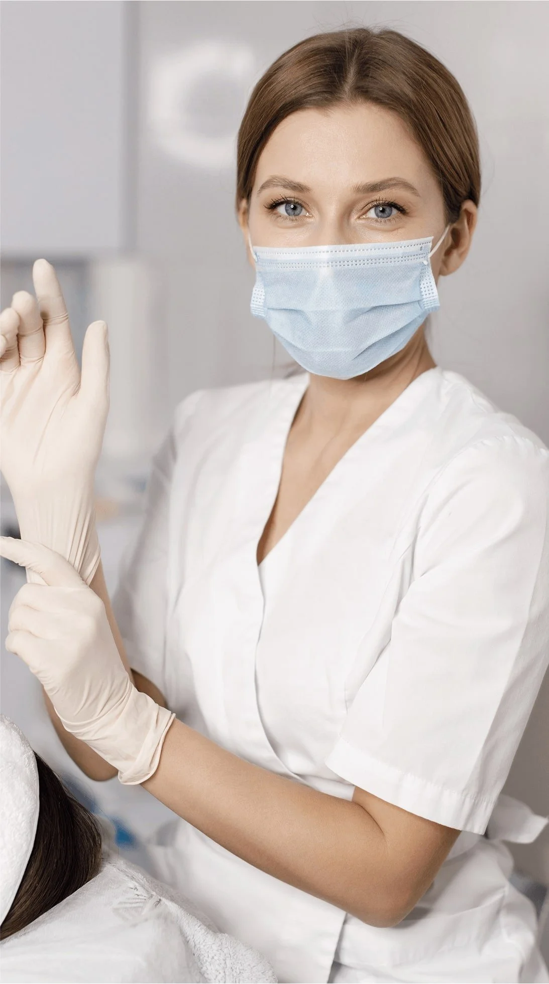 A female medical professional wearing a surgical mask and gloves, preparing to administer an injection or perform a medical procedure.