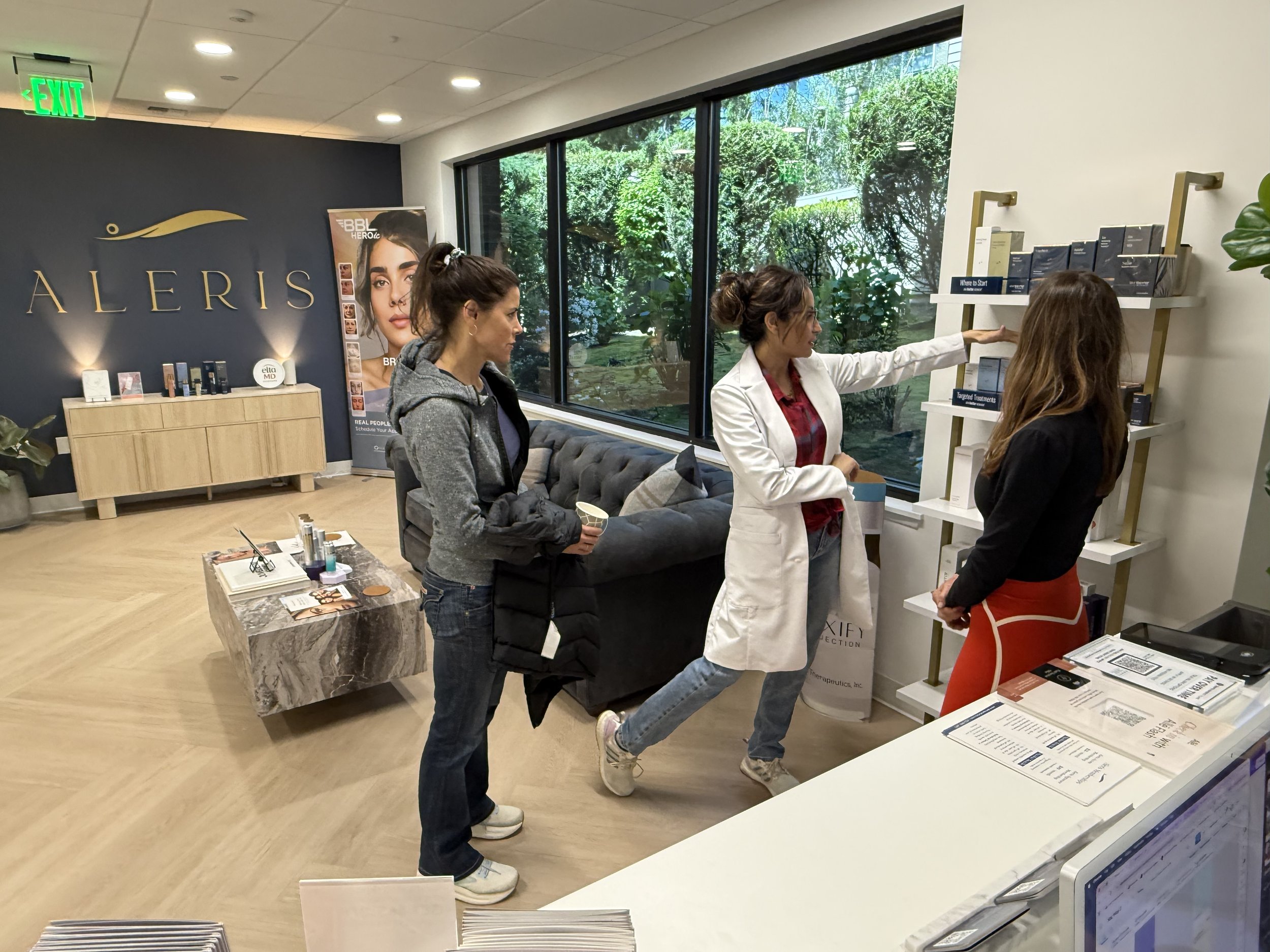 Three women are inside a modern medical or clinic office, with two of them viewing skin care or medical products on a white shelf, and the third woman observing. The interior has large windows with a view of greenery outside, a dark accent wall with a gold logo, and a wooden cabinet with products and promotional materials. A marble coffee table with brochures is in the foreground.