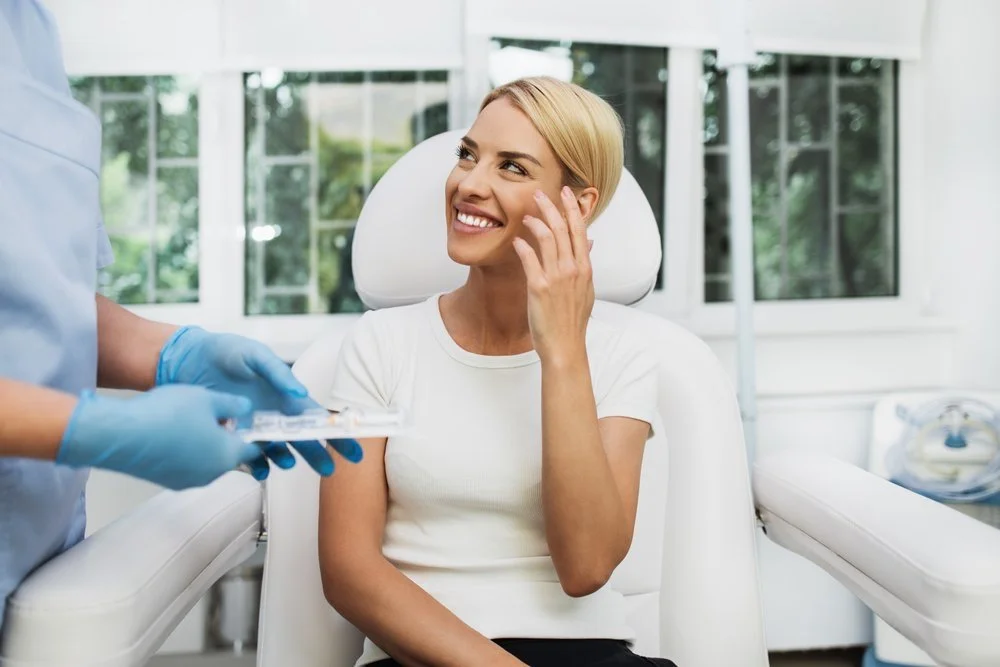 A woman with blonde hair smiling and touching her face while sitting in a dental chair at a dentist office, with a dentist in blue gloves holding dental tools nearby.