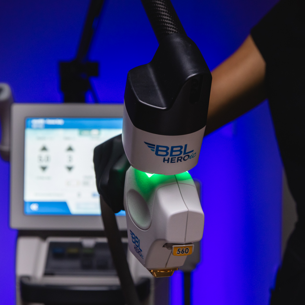 Close-up of a BBL HEROic laser device emitting a green light, with a digital screen in the background displaying settings, against a blue background.