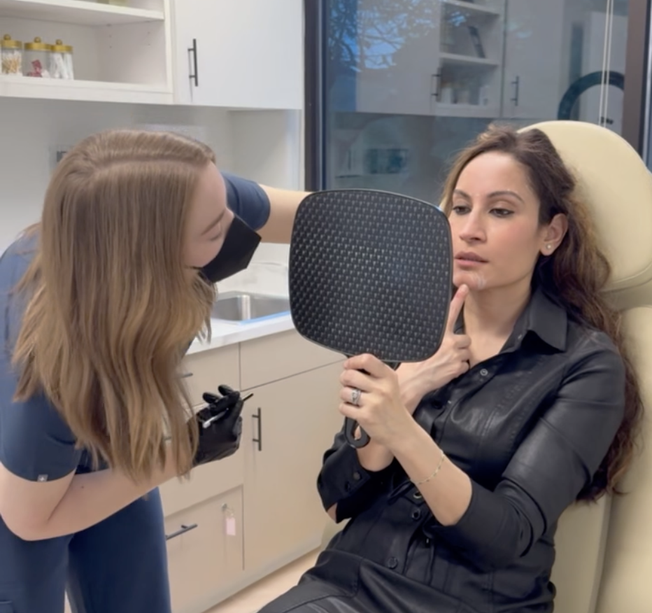 A woman with long brown curly hair sits in a medical office chair, holding a mirror and looking at her reflection. A healthcare professional with blonde hair, wearing a black mask and blue scrubs, is leaning in to examine her face.