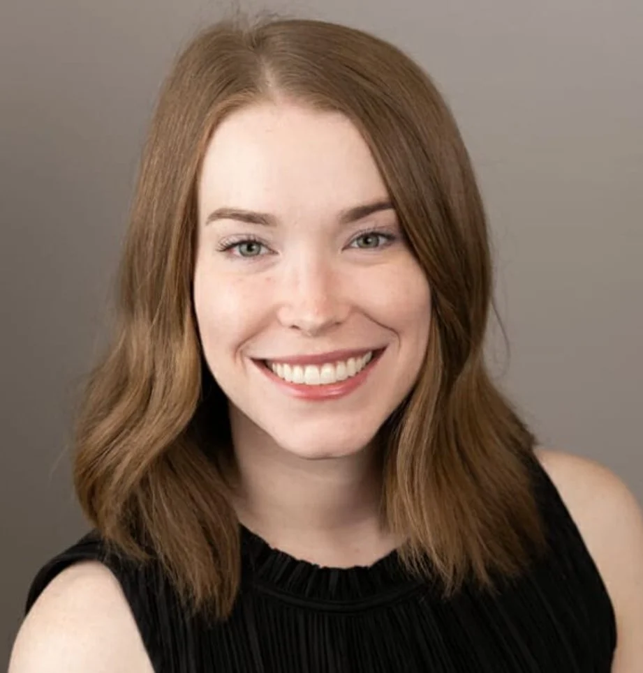 Headshot of a smiling woman with shoulder-length red hair wearing a black sleeveless top.