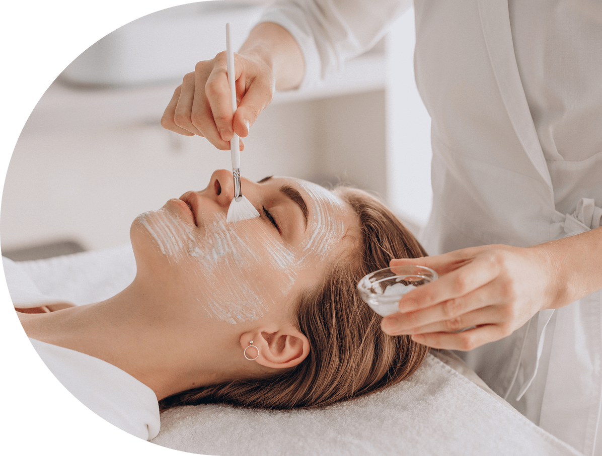 A woman receiving a facial treatment at a spa, with a skincare professional applying facial mask with a brush and holding a small glass bowl.