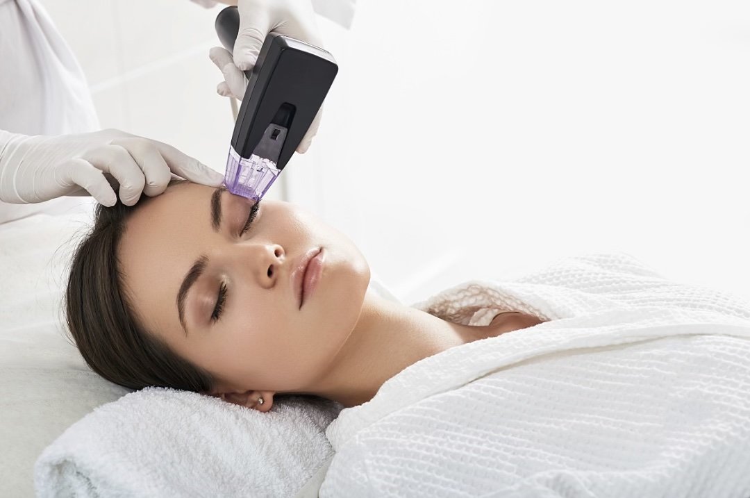 A woman receiving a cosmetic treatment with a handheld device on her forehead, lying on a bed with white sheets, in a clinical setting.