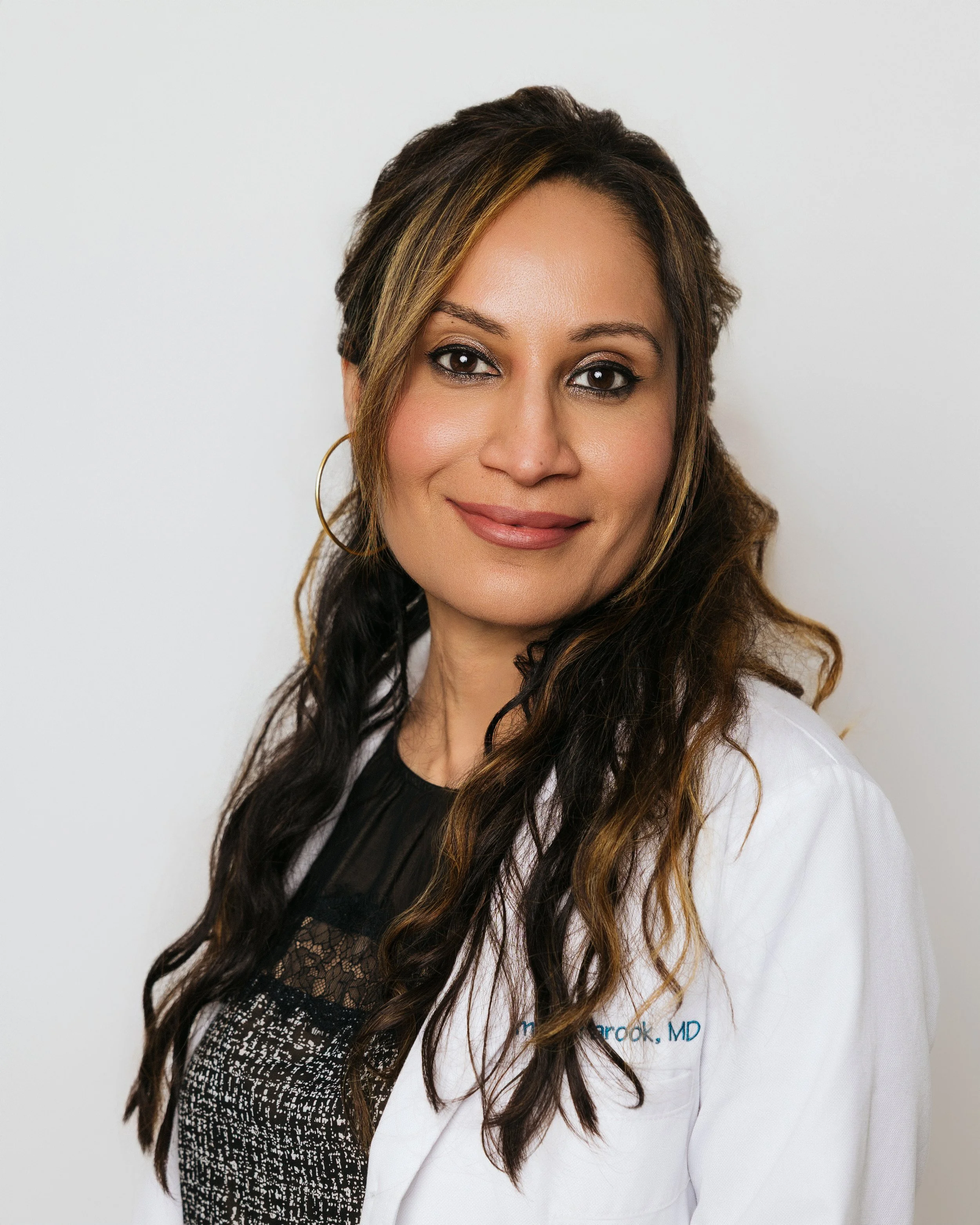 Portrait of a woman with long brown hair and blonde highlights, wearing a white coat and black lace top, smiling against a plain white background.