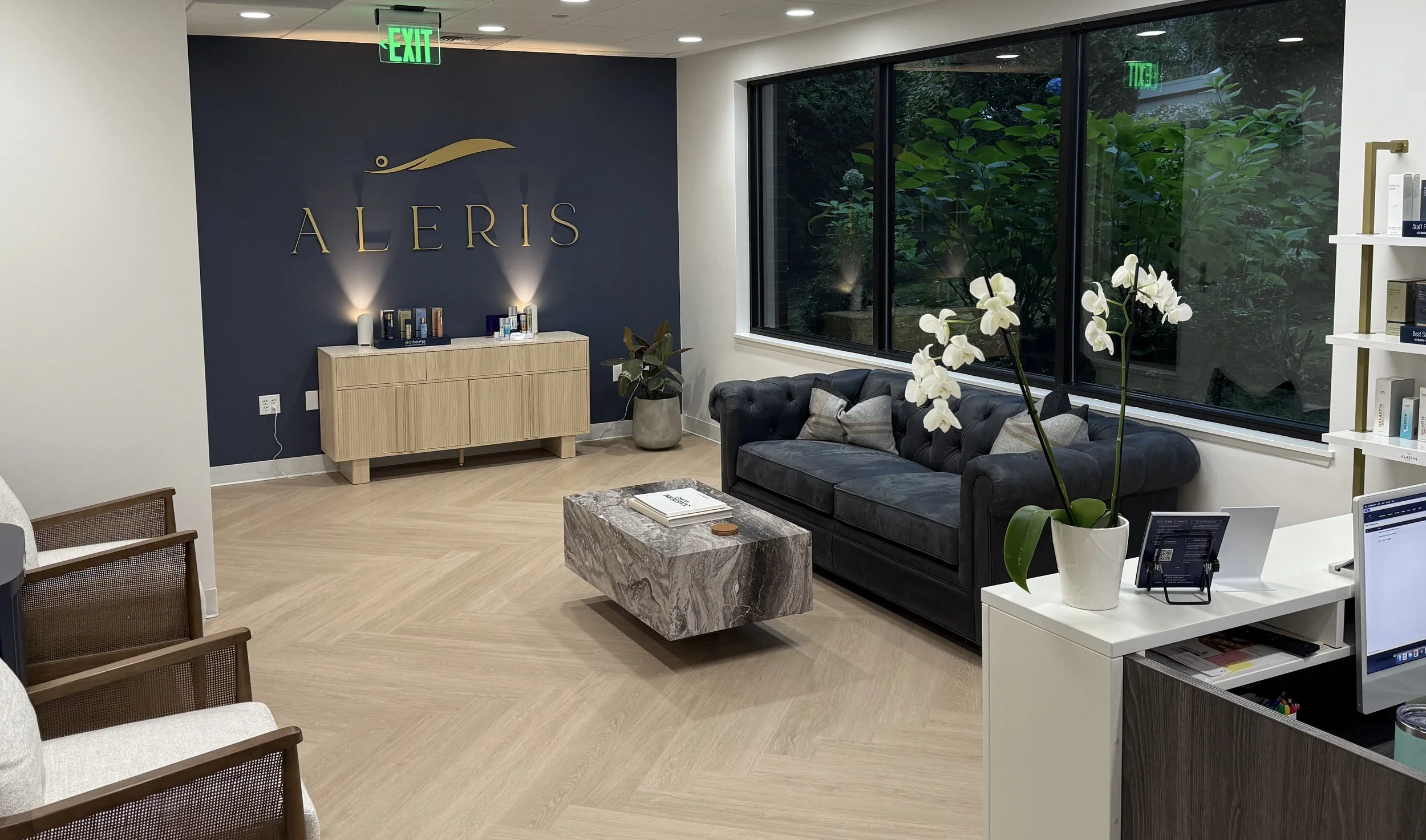 Modern office reception area with a navy wall, gold logo reading 'ALERIS', a black leather sofa, white orchids on a white desk, large window with greenery outside, and a marble coffee table.