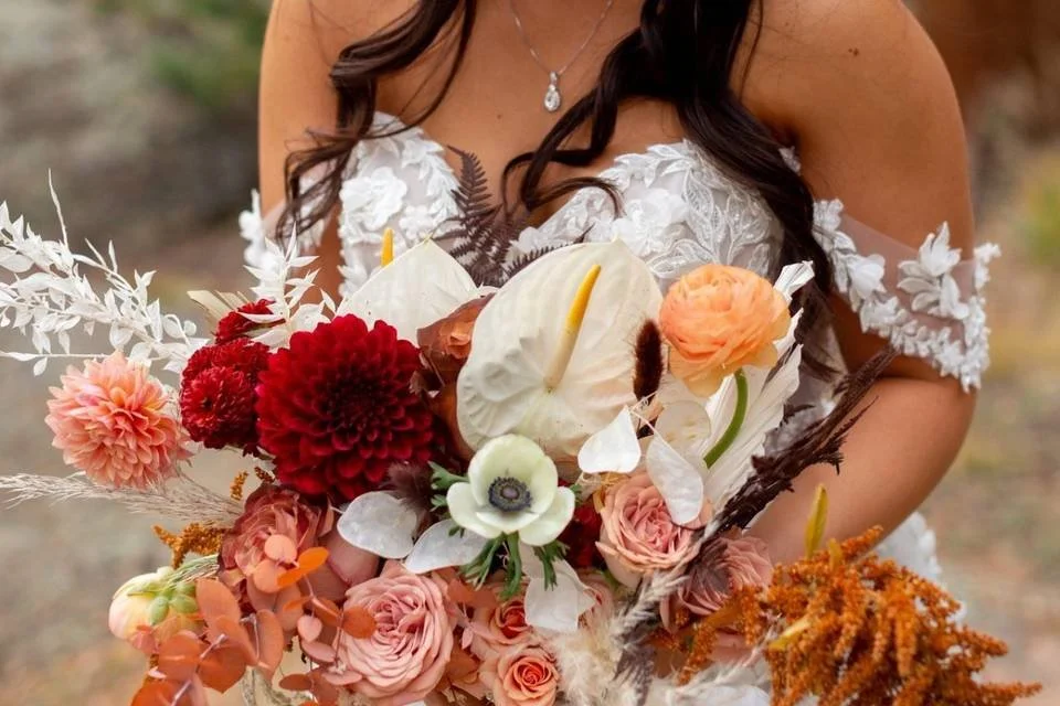 A woman in a white lace wedding dress holding a large bouquet of flowers in shades of pink, red, cream, and orange, including roses, dahlias, and anemones.