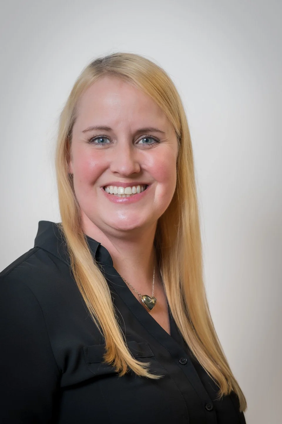 A smiling woman with blonde hair and blue eyes, wearing a black collared shirt and a necklace with a heart-shaped pendant, posing against a plain white background.