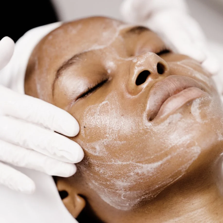 A person receiving a facial treatment with a white facial mask applied, lying down with eyes closed while a gloved hand gently touches their face.