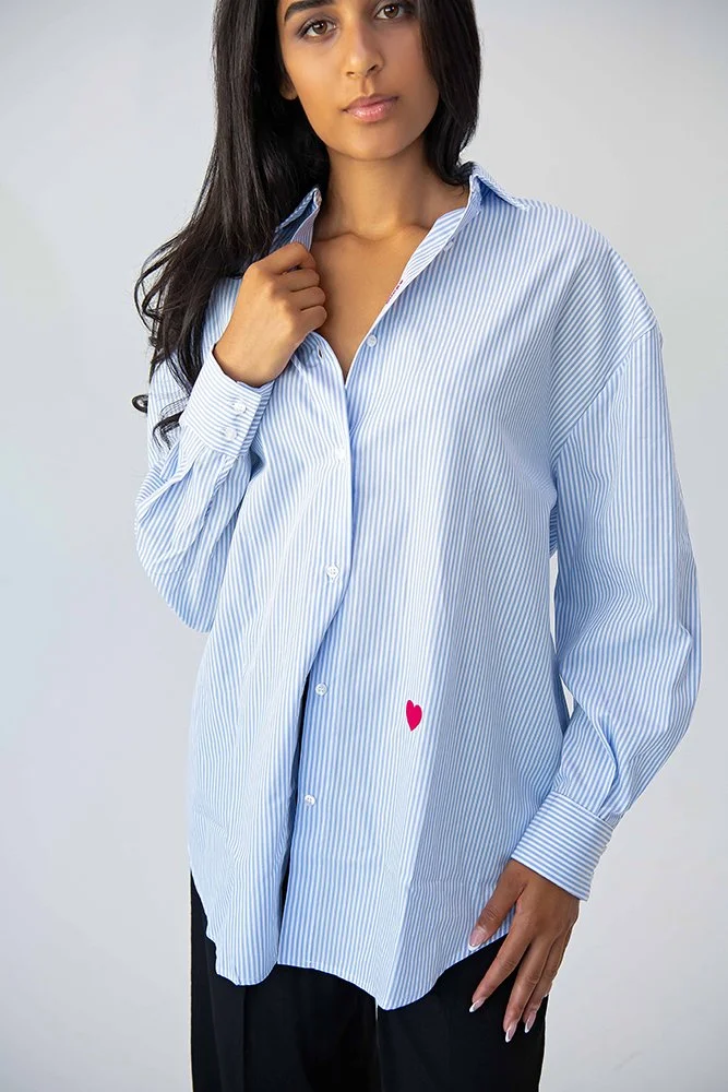 Modell trägt ein hellblau-weiss gestreiftes, oversized Unisex-Hemd mit pinkem Herz-Detail. Made in Italy, designed in Zürich.